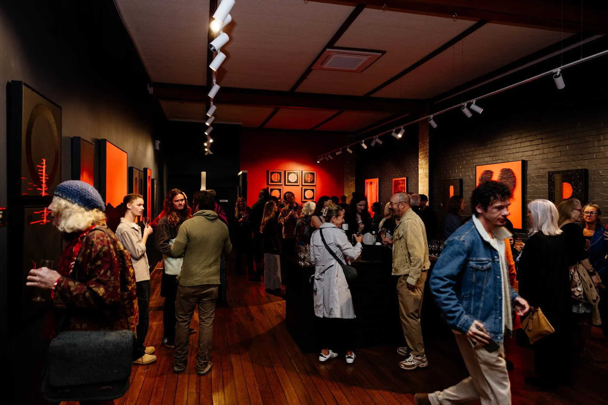 People gathered at an art gallery exhibition, viewing artwork on dark-colored walls with concrete and brick accents. The room has wooden floors, ceiling-mounted track lighting, and bright, colorful artwork displayed.
