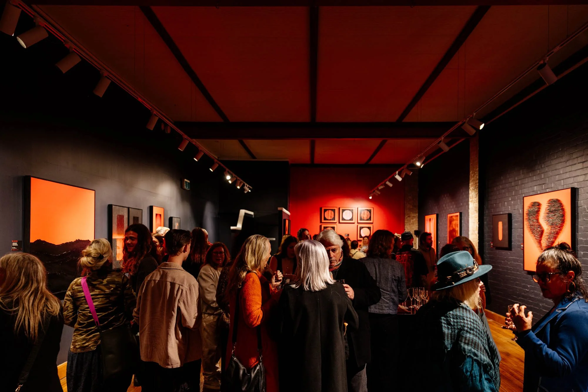 People gathered in an art gallery with red lighting, black walls, and various framed artworks displayed, including an abstract portrait. The scene is lively with glasses of wine.