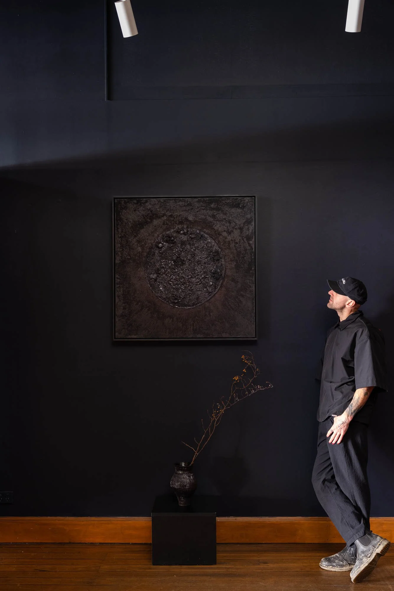 A young man in a black outfit and cap leaning against a black wall in an art gallery. There is a dark square sculpture on a pedestal, a painting on the wall, a vase with a branch, and ceiling lights in the room.