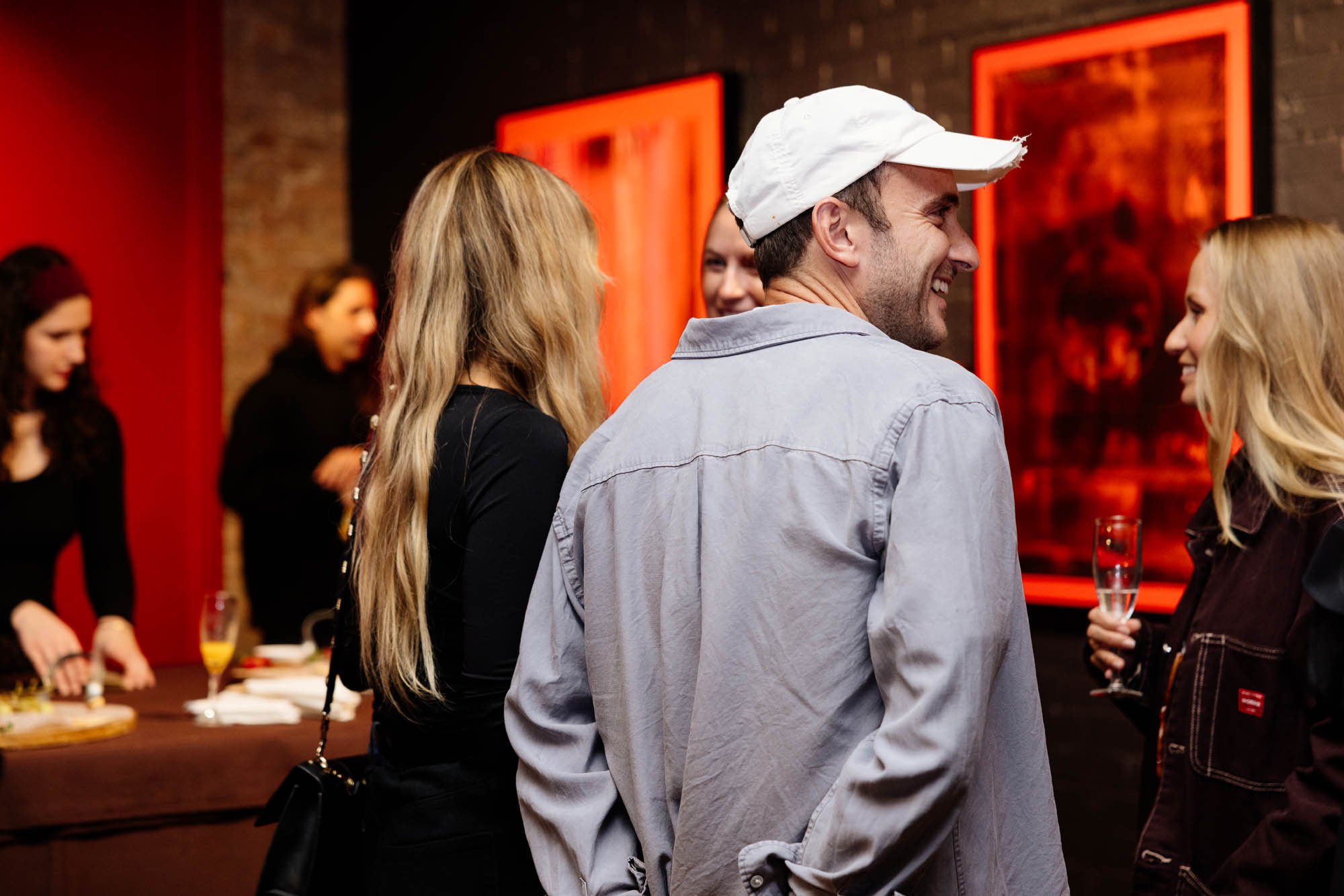 Group of people socializing at a bar or restaurant, including a smiling man wearing a white cap and gray jacket, and women with long blonde hair holding glasses of drinks.