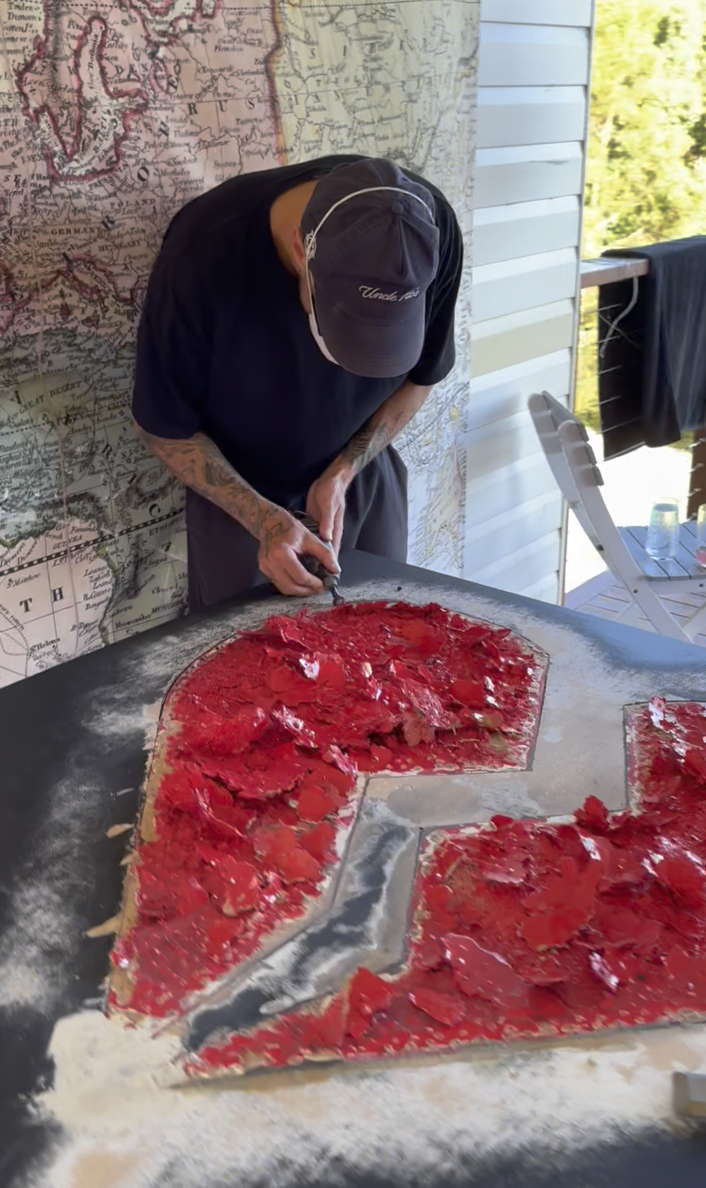 Person in a black shirt and gray cap creating abstract art on a large canvas with red and black paint inside a bright outdoor space.
