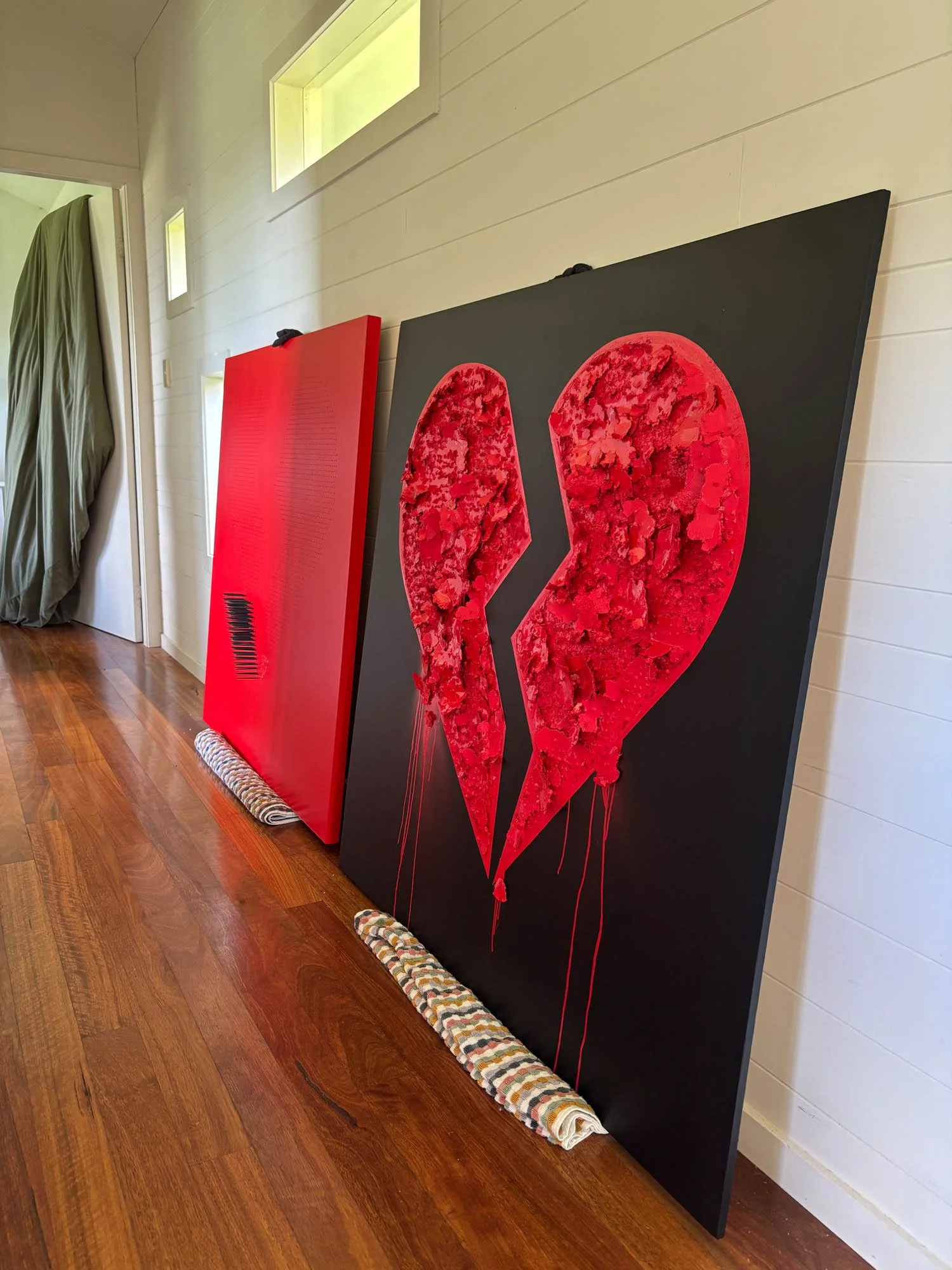 Two paintings of a broken heart, one with a black background and the other with a red background, leaning against a wall. The paintings show a heart with a jagged crack down the middle.