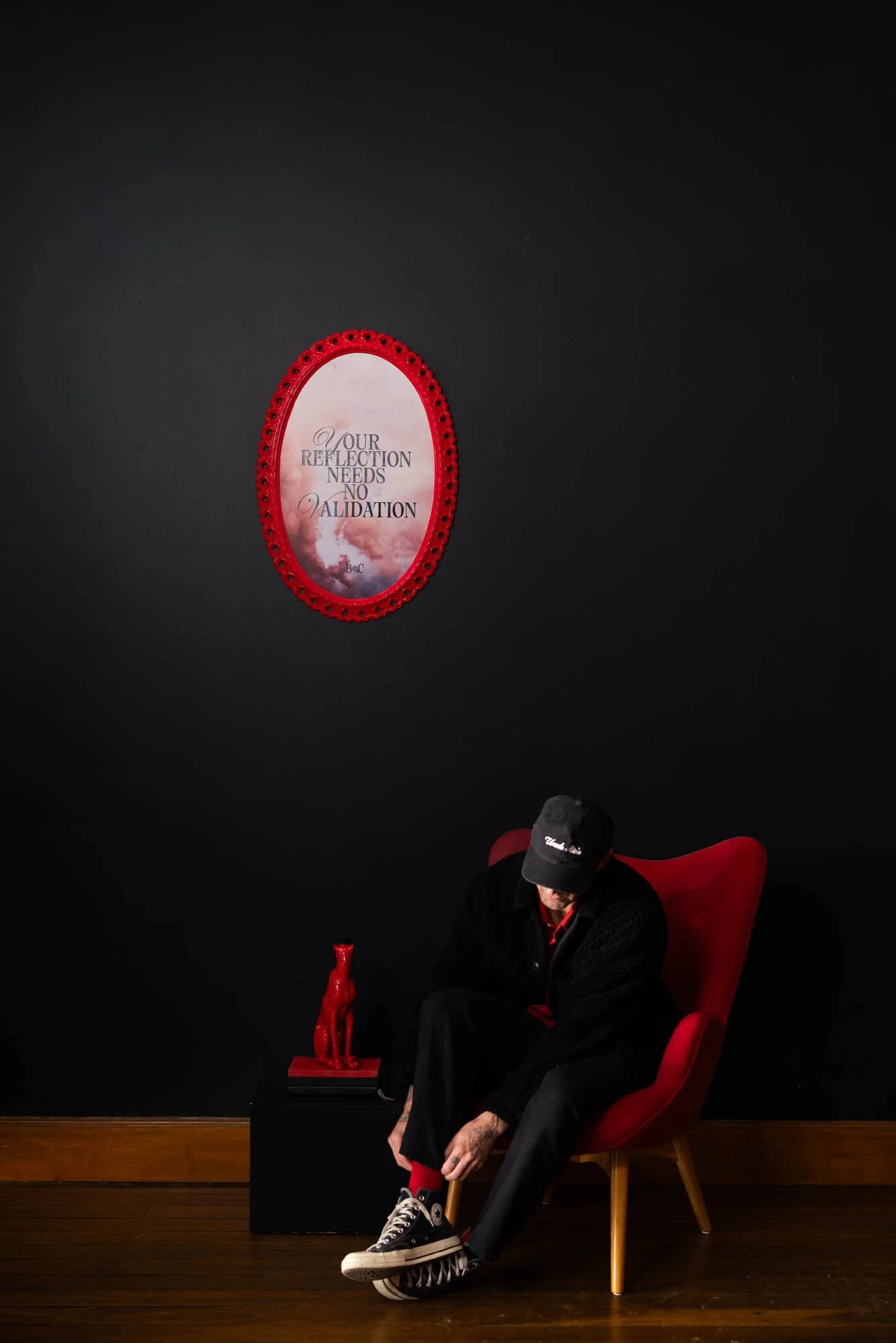 A man sitting in a red chair, adjusting his sock. He is dressed in black with a red shirt underneath. The background is dark, with a red framed mirror and sculpture on a black stand behind him. The floor is wooden.