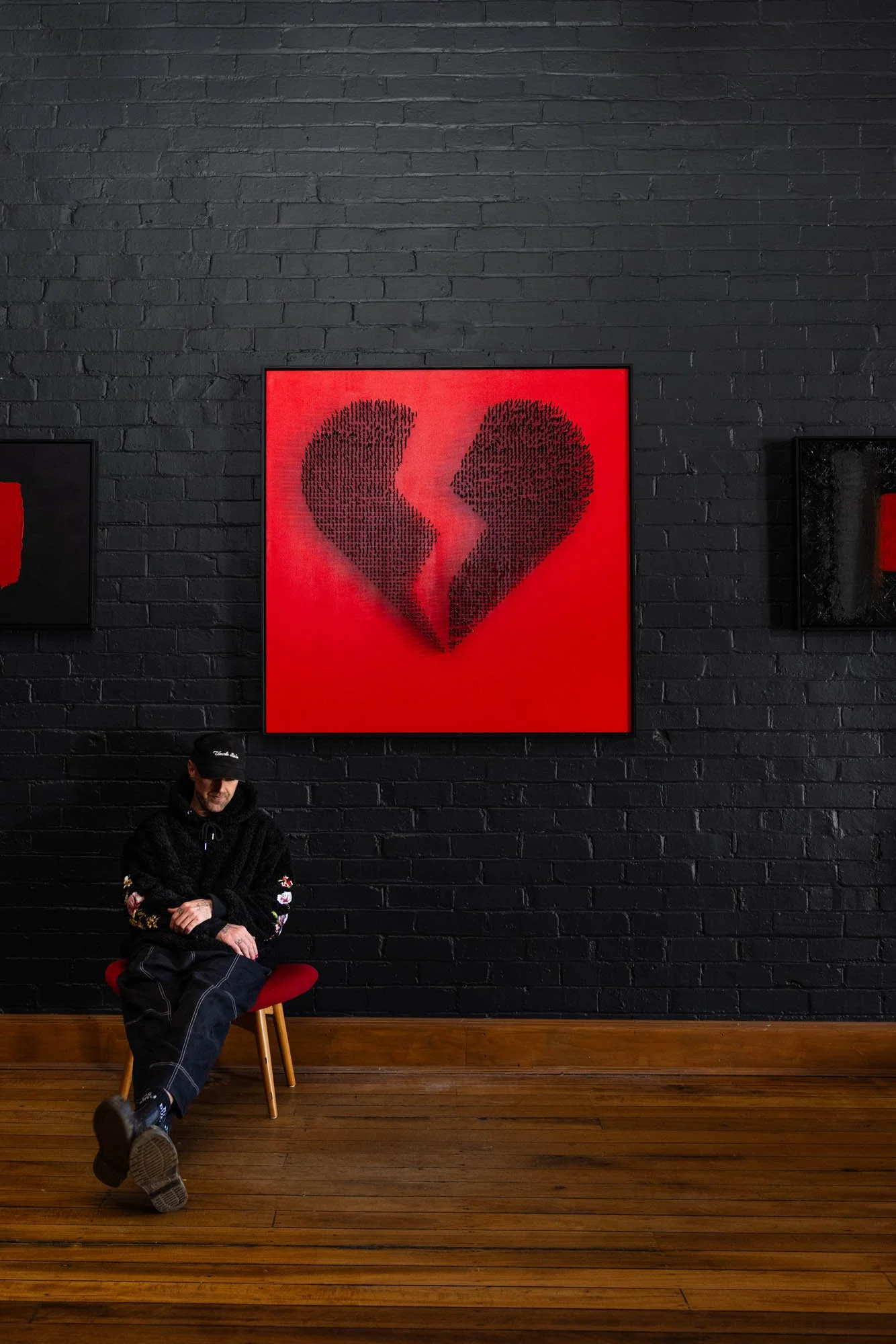 A person sitting on a red stool with a black jacket and dark jeans, head down and arms crossed, in front of a black brick wall with three art pieces, including a red canvas with a black broken heart design, and a wooden floor.