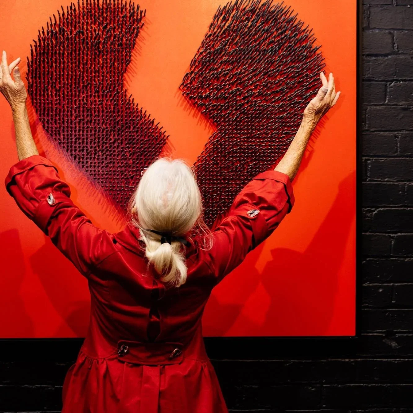 An elderly woman with white hair in a ponytail, wearing a red jacket, is standing with her back to the camera, raising her arms to touch a large red artwork on a wall. The artwork depicts a broken heart shape, with numerous small objects creating a textured surface.