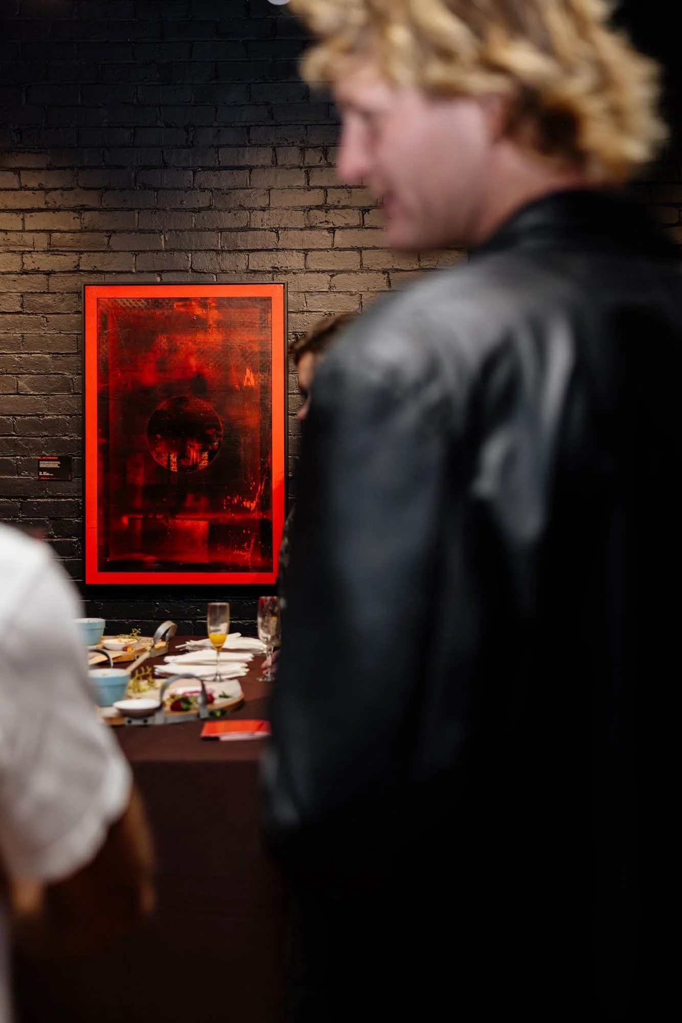 People gathered around a table with food and drinks at an art gallery with a dark brick wall and red artwork in the background.