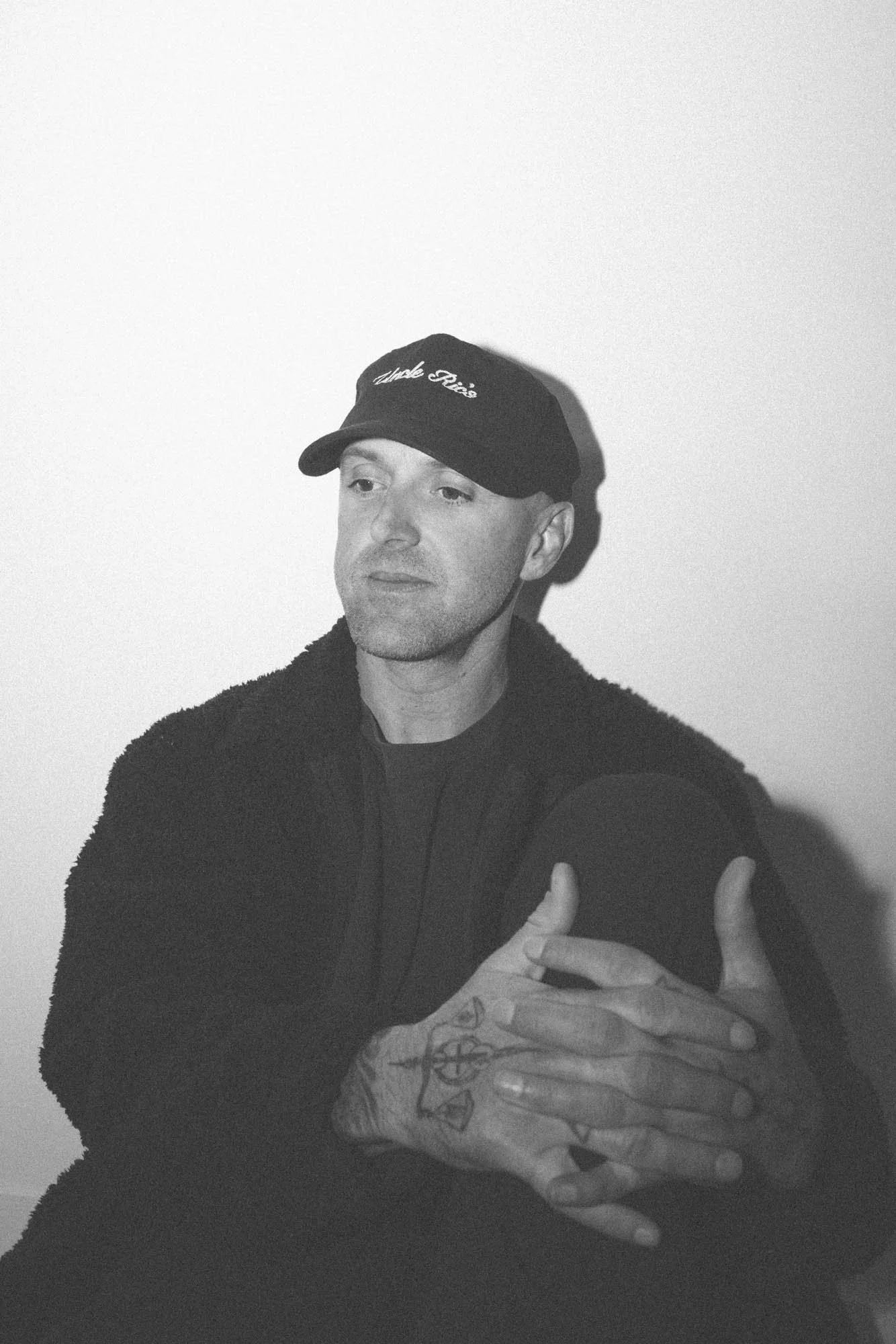 A man wearing a black cap with white text, a black jacket, and tattoos on his hand, making a gesture with his hand.