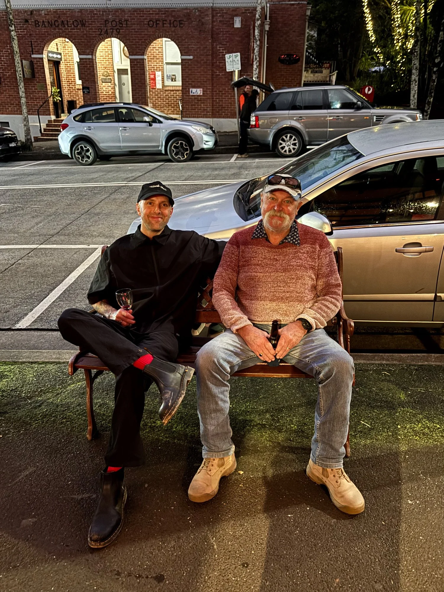 Two men sitting on a bench at night, both smiling, with one holding a glass of wine and the other holding a beer. Behind them is a silver car, and in the background, there are parked cars, a brick building with an entrance, and trees with string lights.