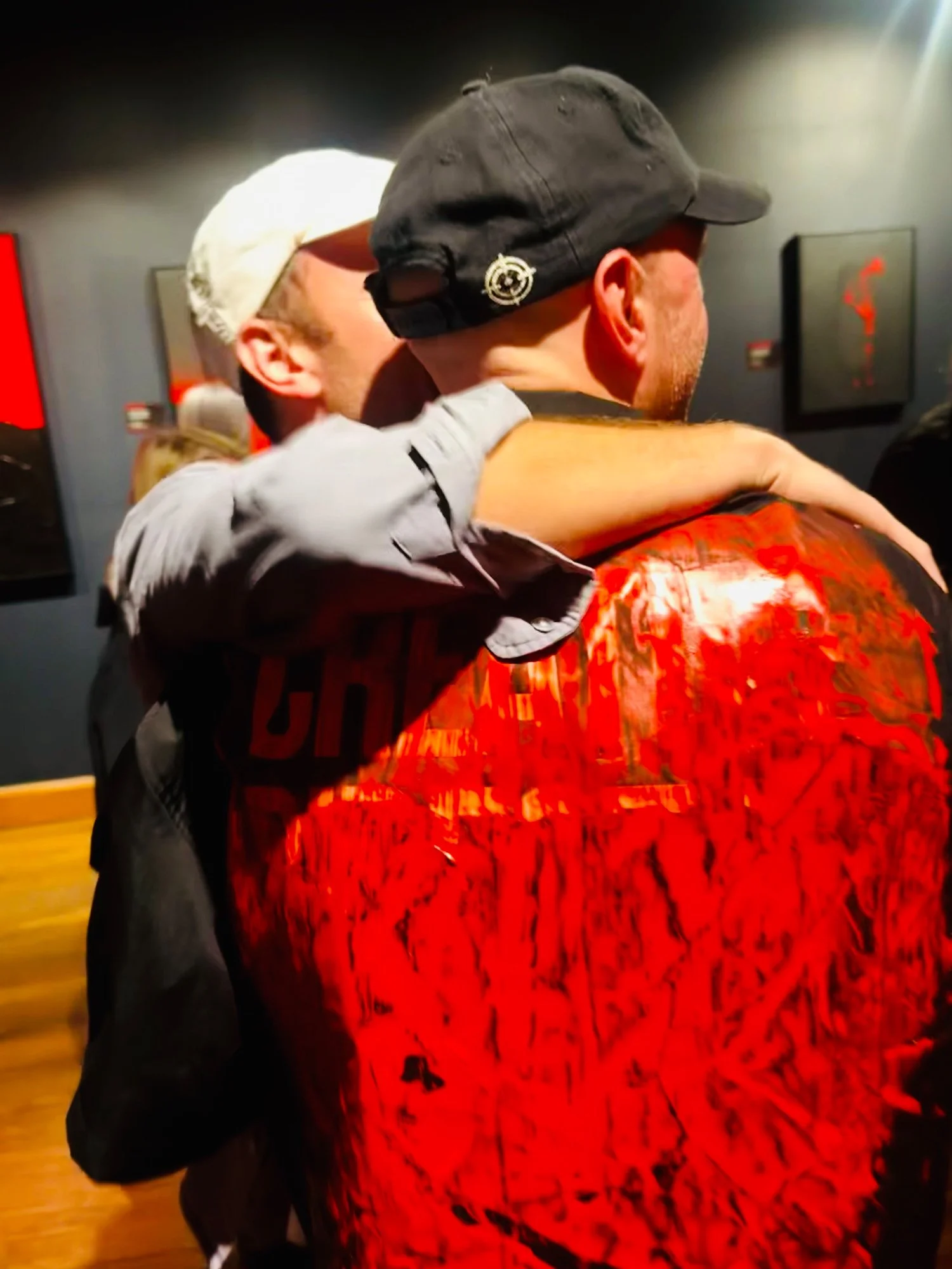 Two men hugging each other closely, one wearing a black cap and the other wearing a white cap, with a dark wall and framed art in the background.