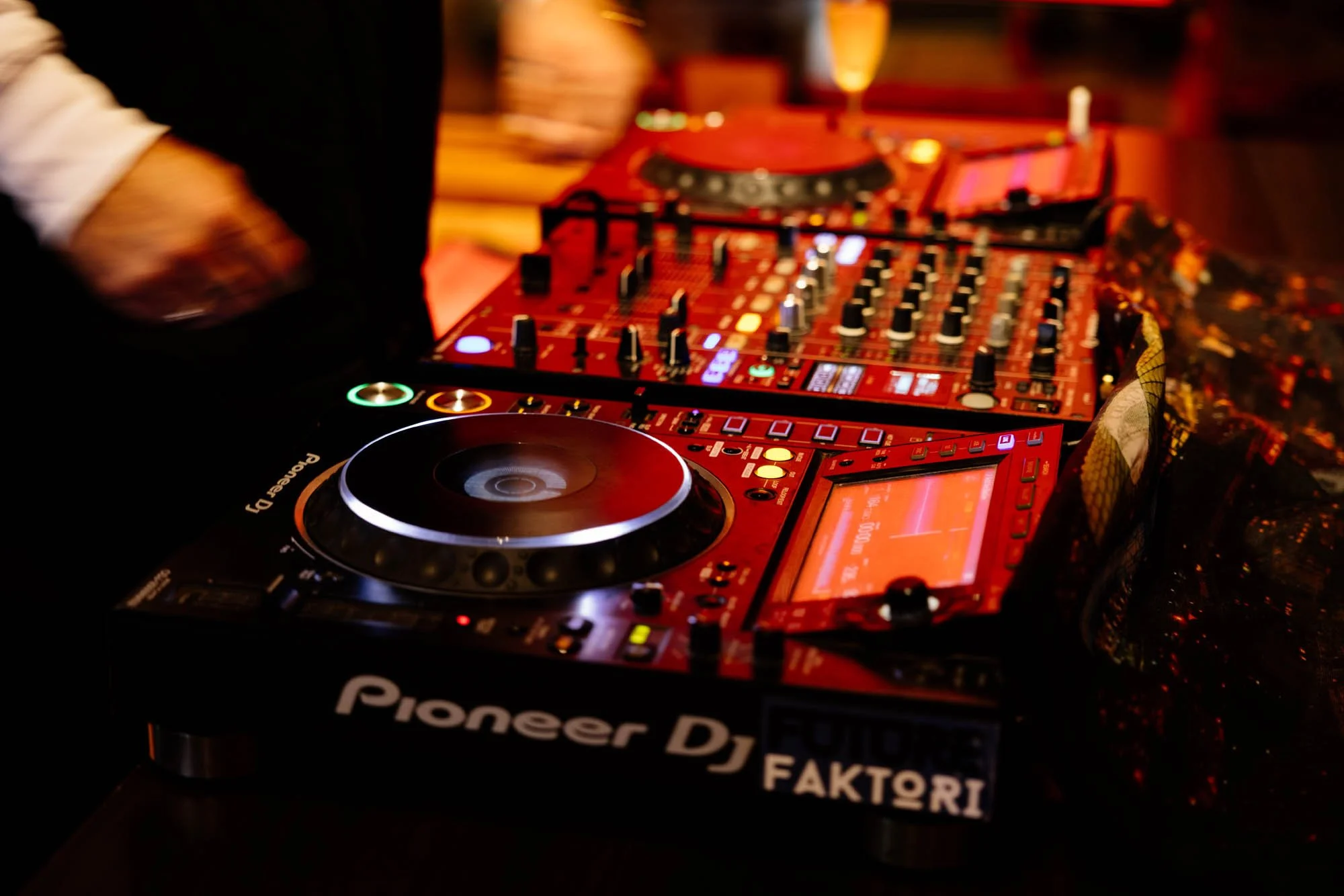 A DJ mixing music on Pioneer DJ equipment in a dimly lit setting with warm, orange and red lighting.