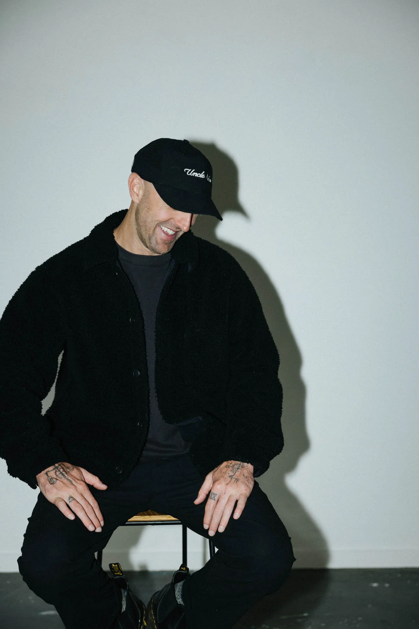 A man with tattoos on his fingers sitting on a stool, wearing a black cap, black sweater, black pants, and black boots, against a plain gray wall, smiling and looking down.