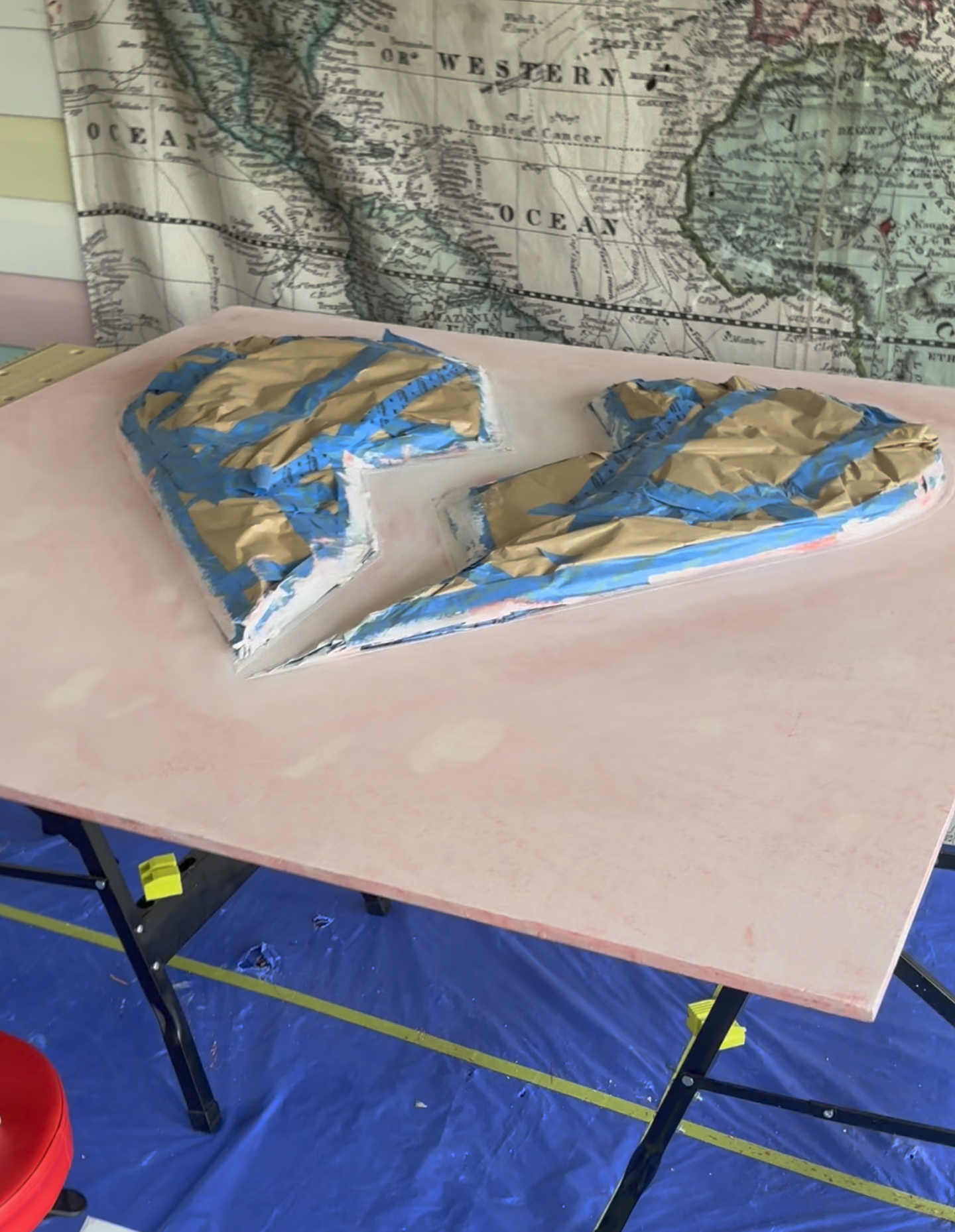 A table covered with pink paper, with two crumpled paper shapes wrapped in brown paper and blue tape on top, set against a backdrop of a map of the Western United States.
