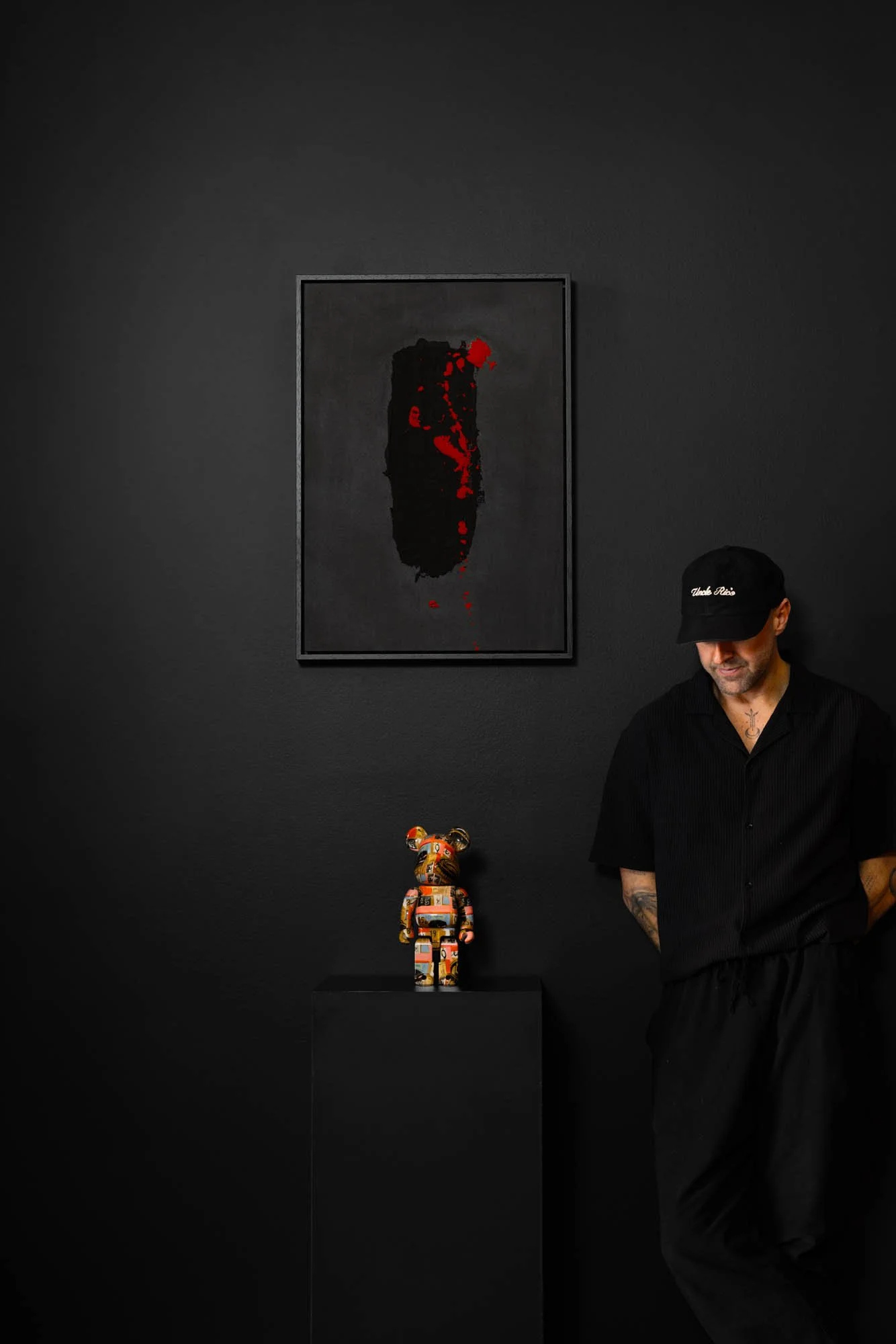 Man wearing black clothing, a black cap, and arm tattoos, standing next to a colorful sculpture of a bear on a black pedestal. Behind him is a dark wall with a framed abstract black and red art piece.