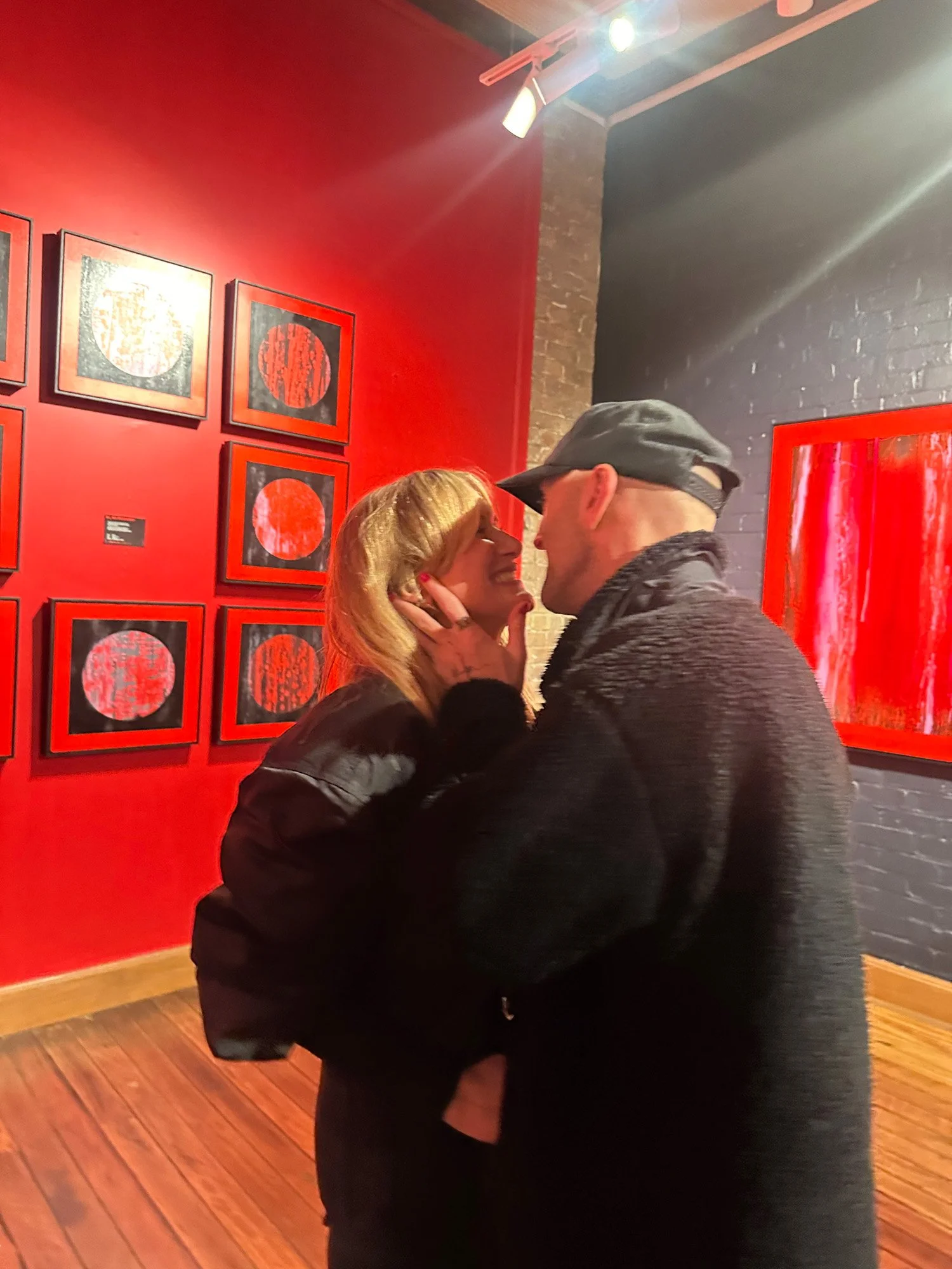 A couple standing close and smiling at each other in an art gallery with red walls and framed artworks, while the woman touches the man's face.