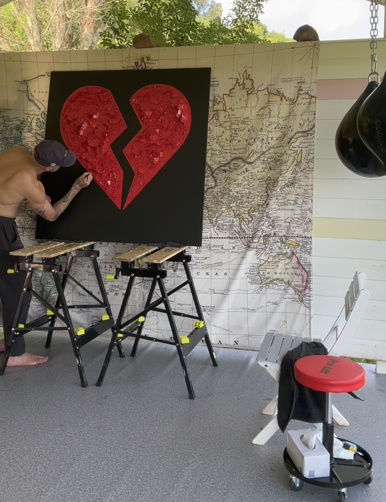 Person working on a large painting of a broken red heart on a black background, with a large world map mural on the wall behind them and a window with trees outside. There is a white bench and a red stool with a box of tissues nearby.