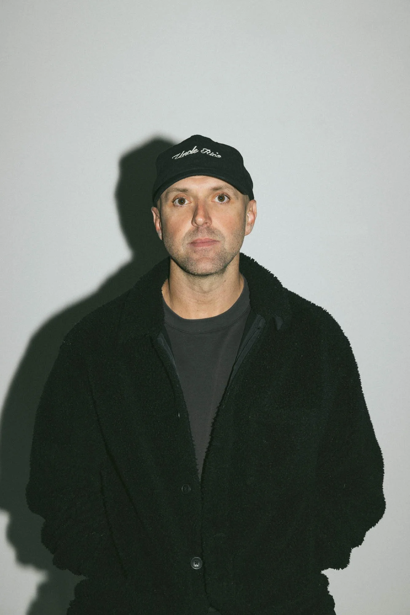A man wearing a black cap with white embroidered text and a black fleece jacket stands against a plain white wall. His shadow is cast on the wall to his left.