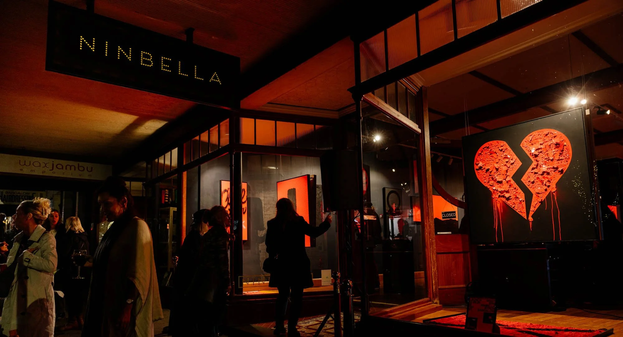 An indoor art exhibit features a large digital display of a broken red heart on the right side, with people observing the artwork. Overhead sign reads 'NINBELLA'.