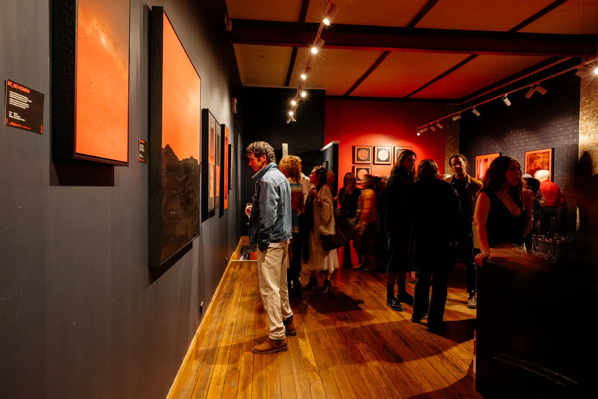 People viewing and discussing artwork in an art gallery with dark walls and wooden floors.