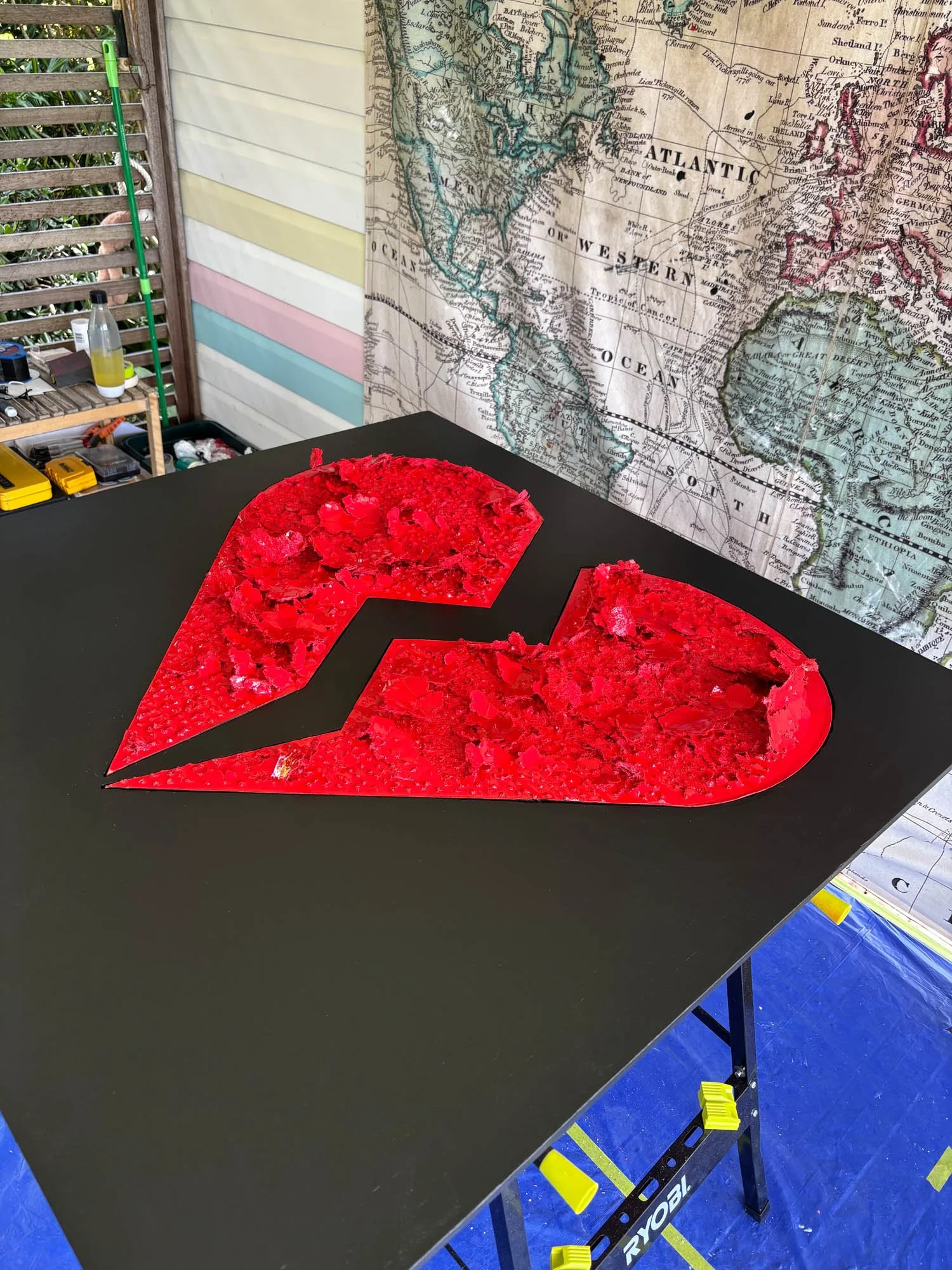 A black table with a red and black graphic design on top, resembling a lightning bolt or a stylized letter 'N'. In the background, there is a world map or vintage map hanging on the wall, part of a cluttered workspace with various tools and bottles on a small wooden shelf, and a green pole leaning against the wall.