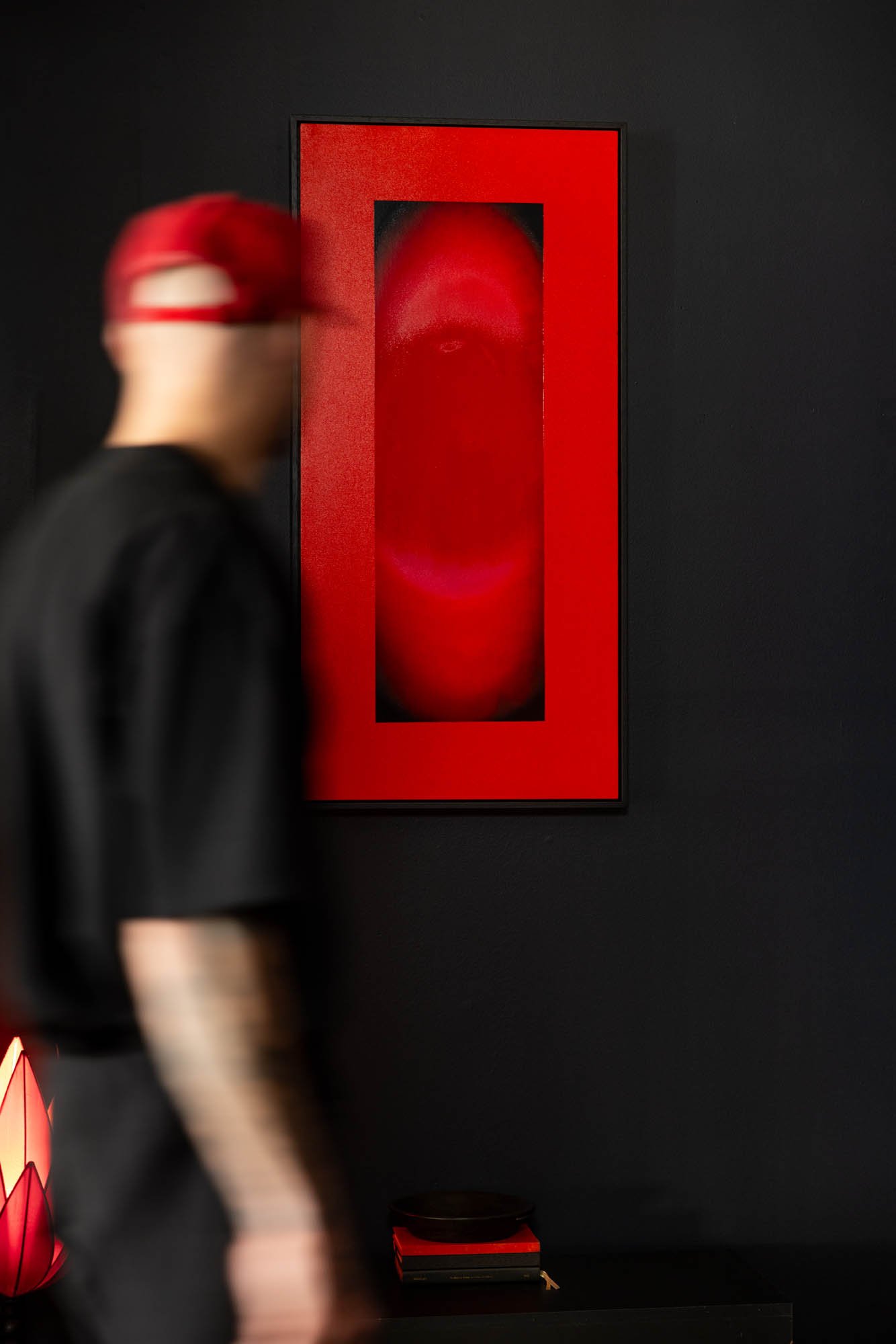 A person wearing a red cap and black shirt stands in front of a black wall with a rectangular red art piece featuring an abstract face in red and black.