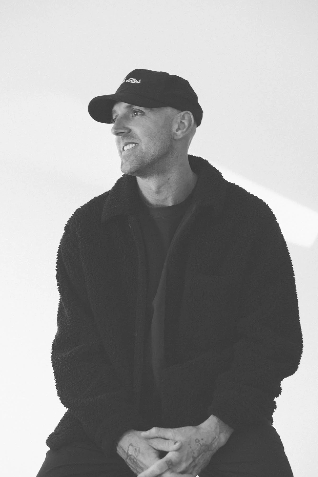 A black-and-white photo of a man in a black cap and black sweater, smiling and looking to the left. He has tattoos on his hands, and his hands are clasped together in his lap. The background is plain and light-colored.
