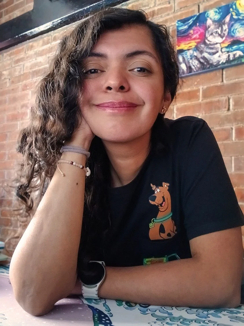 A young woman with curly brown hair, wearing a black t-shirt with Scooby-Doo graphic, sitting at a table in a restaurant with exposed brick wall, smiling and resting her head on her hand.