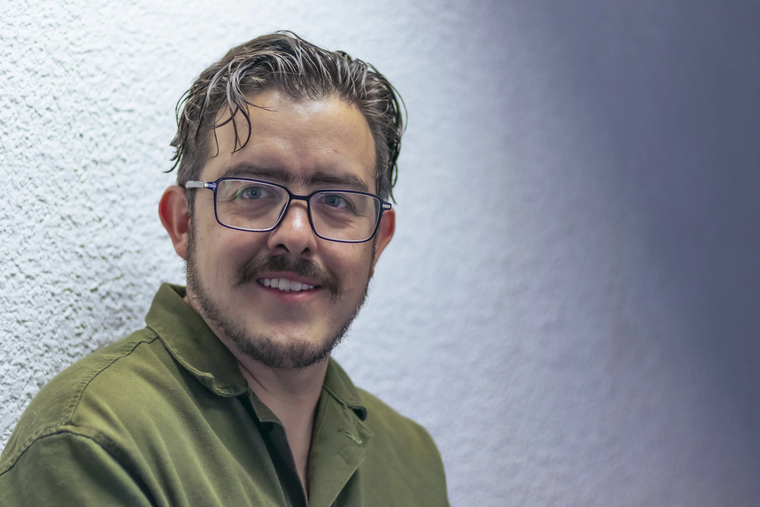 A man with glasses, wavy brown hair, and a beard, wearing an olive green shirt, smiling and standing against a textured wall.