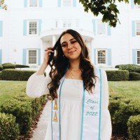 A young woman in a white graduation gown smiling outdoors in front of a white building with greenery.