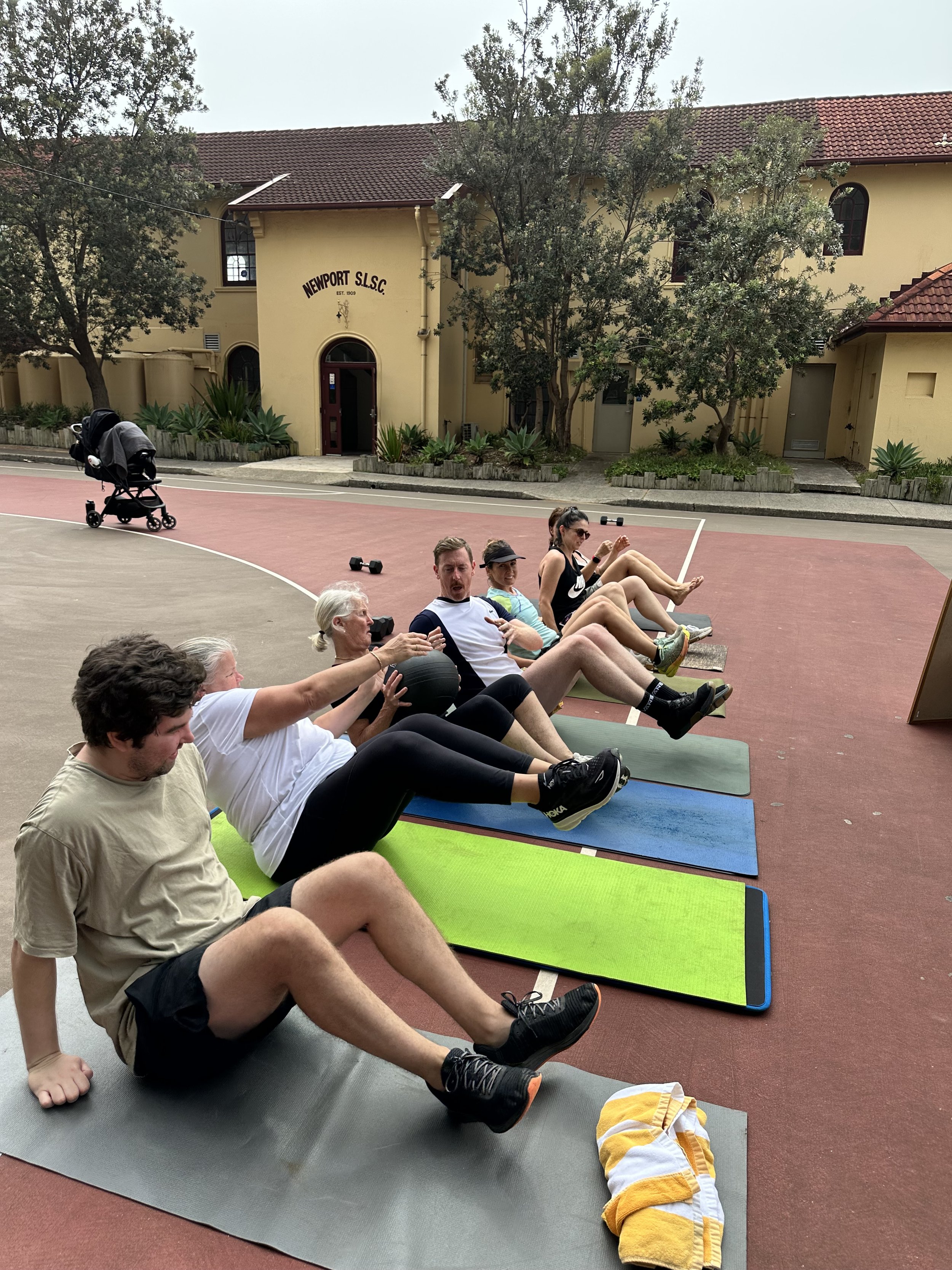 Group of people participating in an outdoor fitness class, doing seated abs exercises on yoga mats, with a pink and gray sports court, and trees and a yellow building in the background.