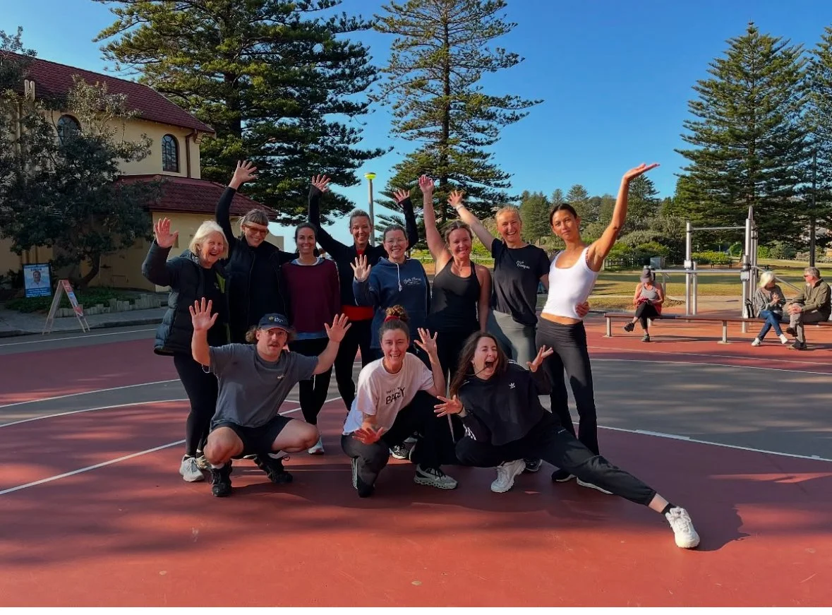 Group training of ten people posing on a red outdoor tennis court with trees and a building in the background, all smiling and raising their hands. Personal training at Newport Beach, outdoor fitness session.