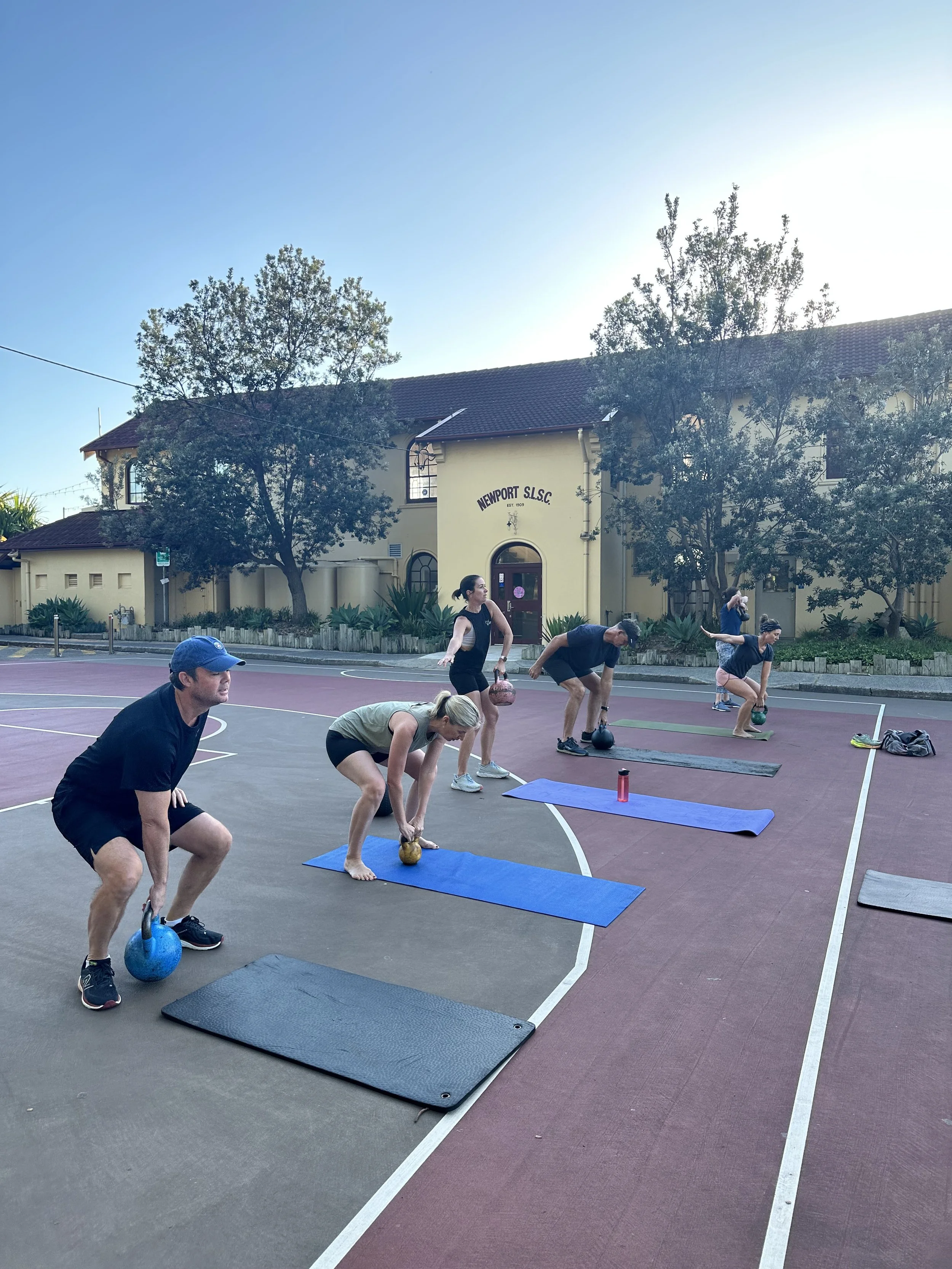 Group of people participating in a kettlebell workout session outdoors on a basketball court, with some using yoga mats.