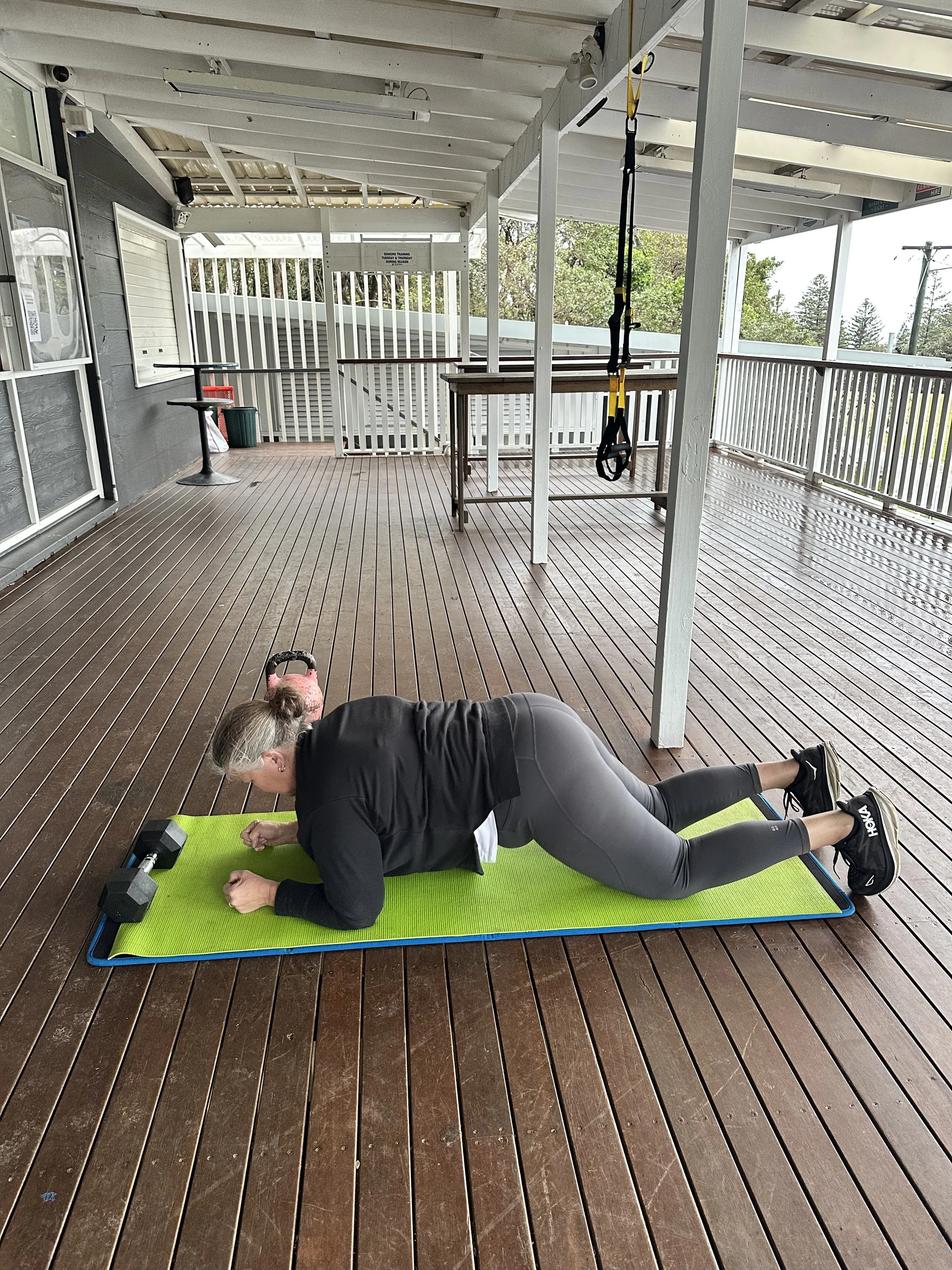 A woman exercises, training outdoors on a wooden deck, using dumbbells and doing a plank exercise. During personal training session.