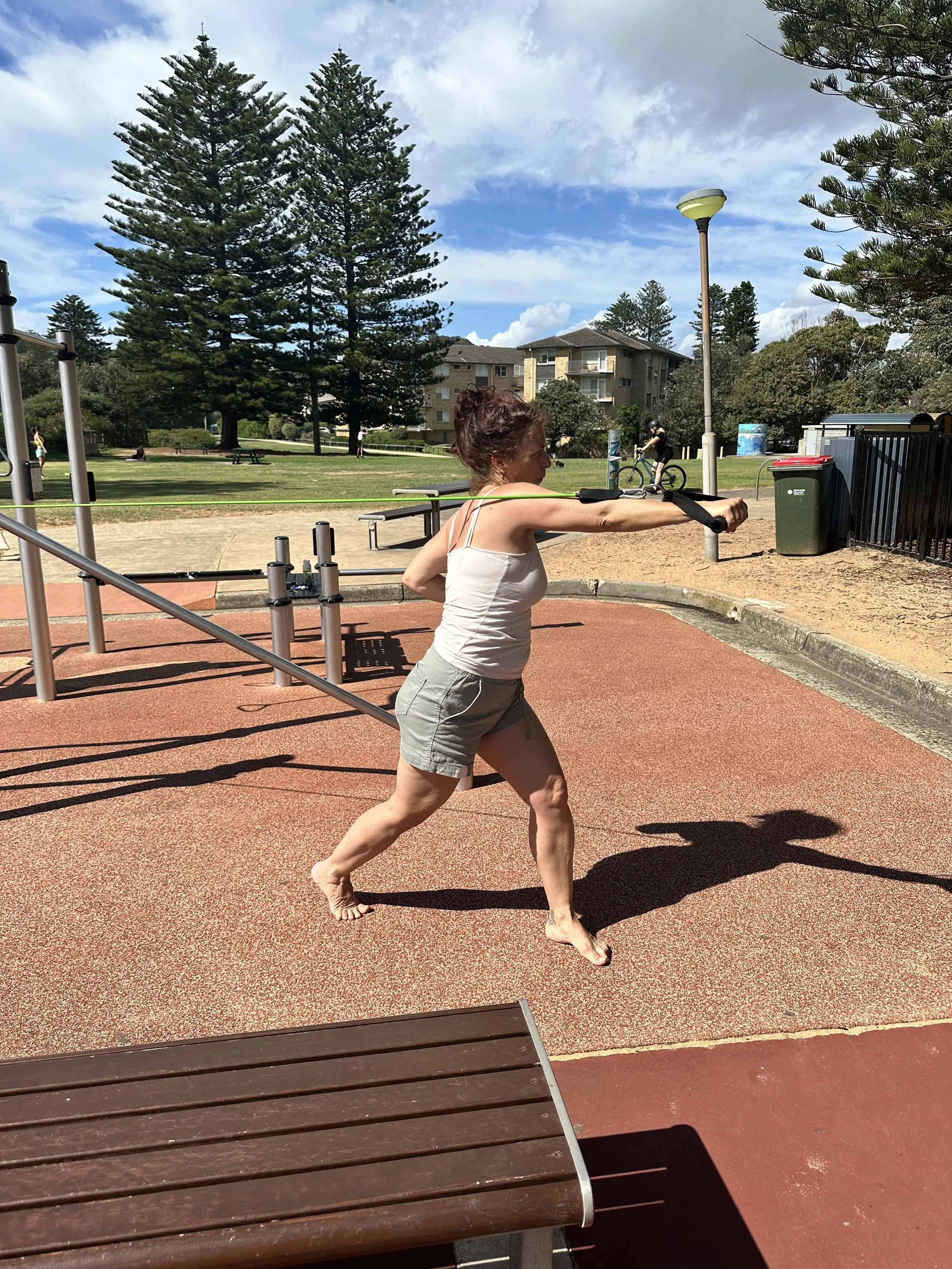 A woman at a playground using a resistance band for exercise, with her arm extended forward and supported by a harness, on a sunny day with a bright blue sky and trees in the background.