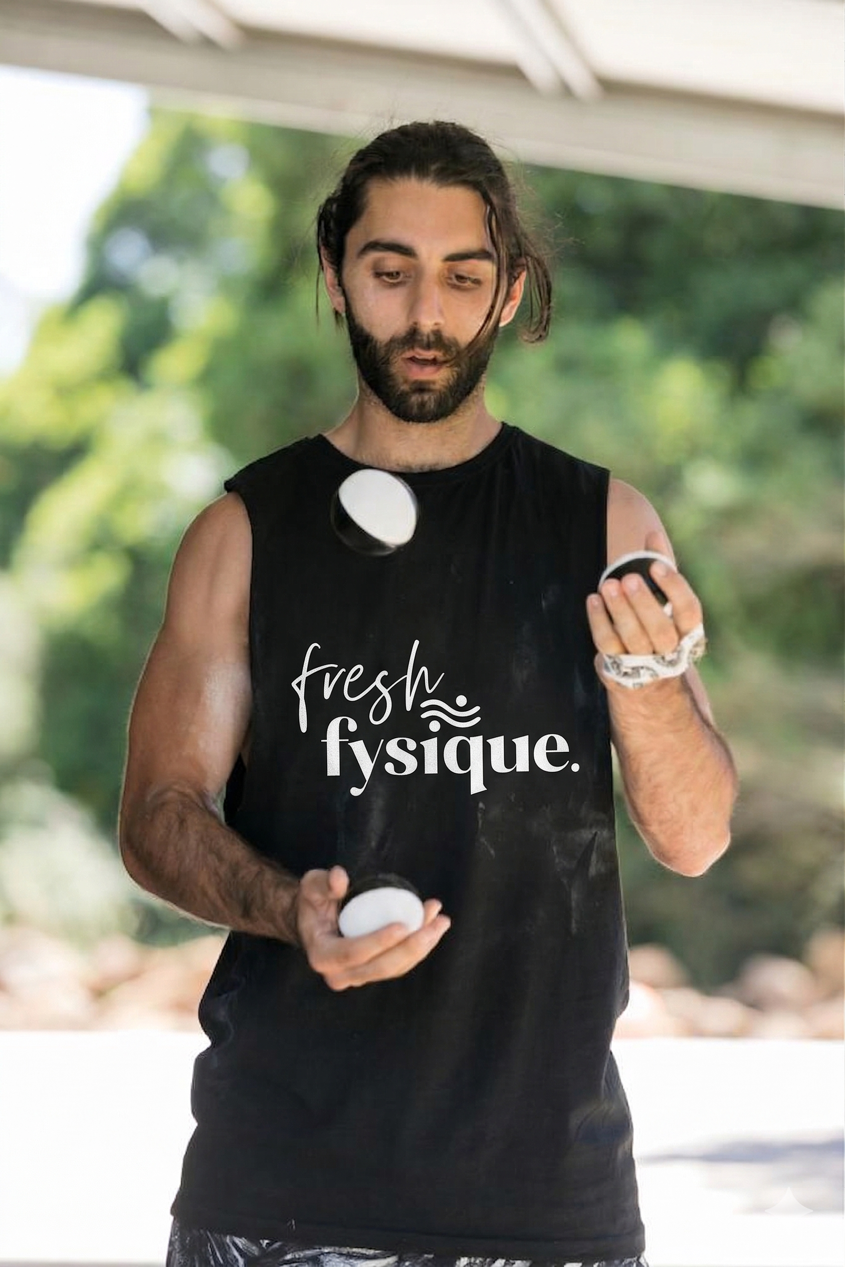 A man wearing a black shirt with the words "fresh physique" on it, is looking at and holding a small container in each hand, outdoors with green trees in the background. Personal trainer at Newport fitness.