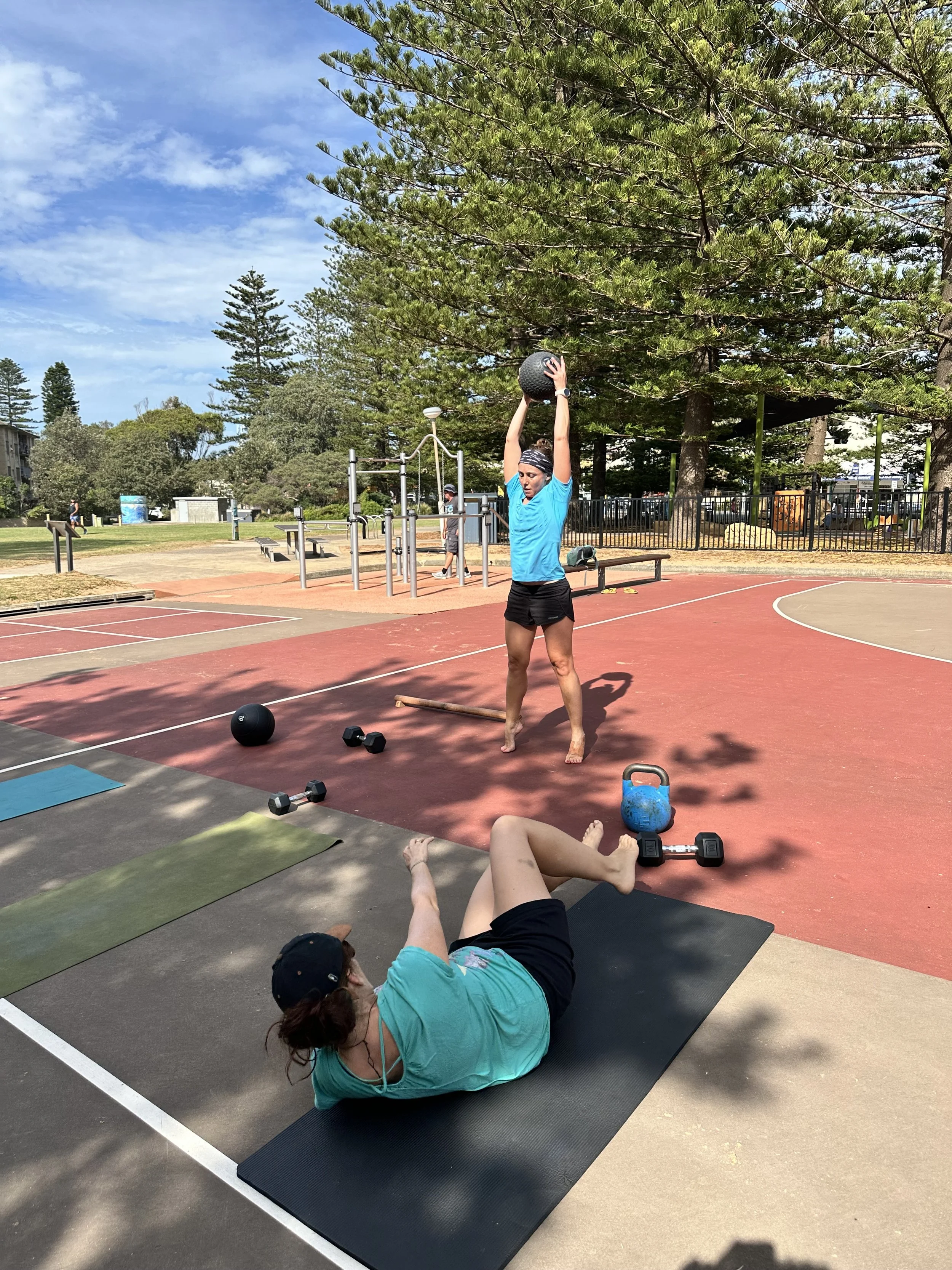 Two women exercising at an outdoor park. One woman is lying on a yoga mat and performing a leg raise exercise, while the other woman is standing and holding a medicine ball above her head. Fitness equipment and large trees are visible in the background.