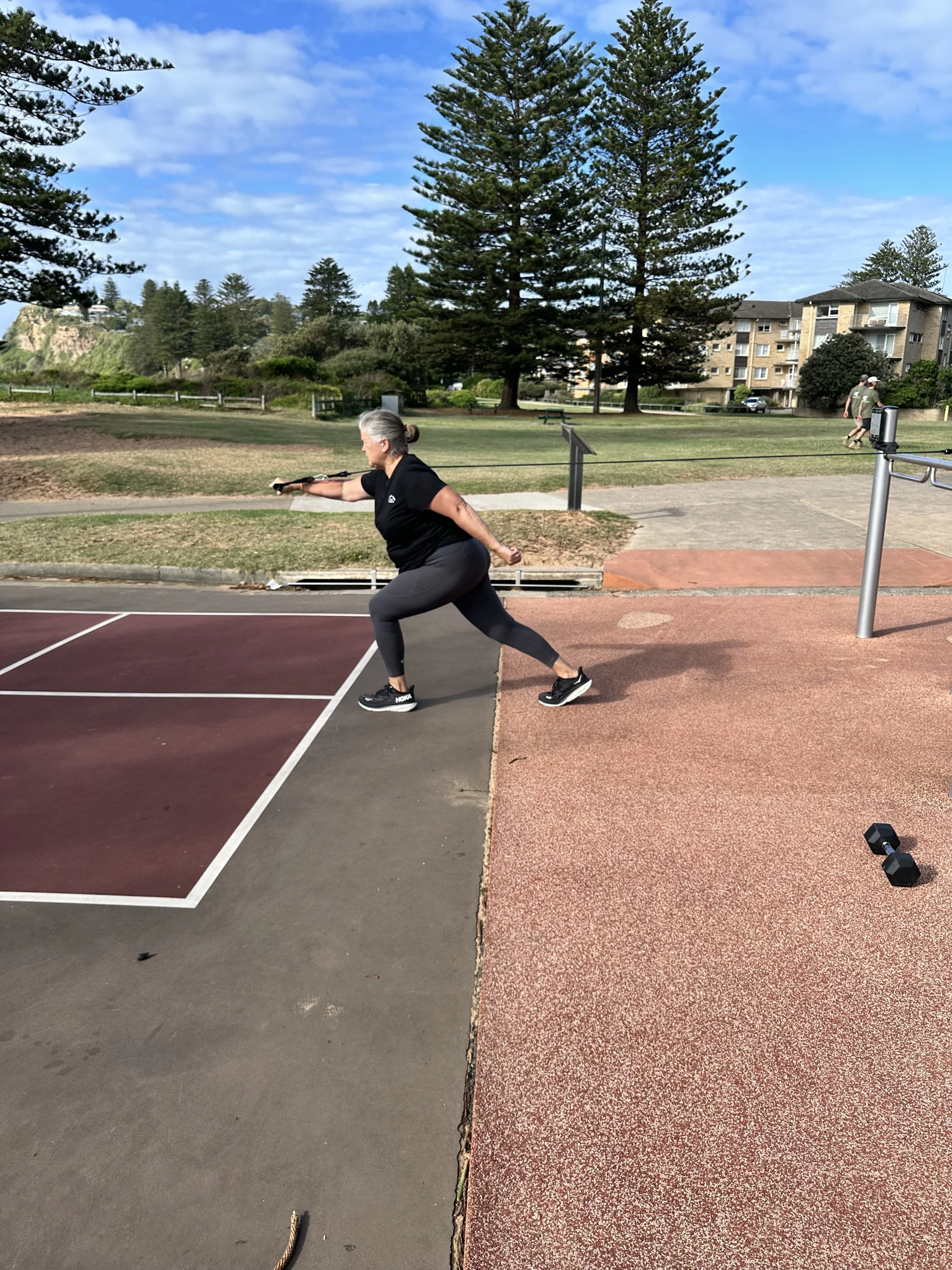 A woman performing a resistance band stretch or exercise outdoors on a playground court, with trees and residential buildings in the background.