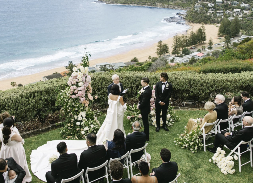 A beach wedding ceremony at Jonah's on a grassy hillside overlooking Whale Beach, with guests seated and a couple exchanging vows with an officiant.