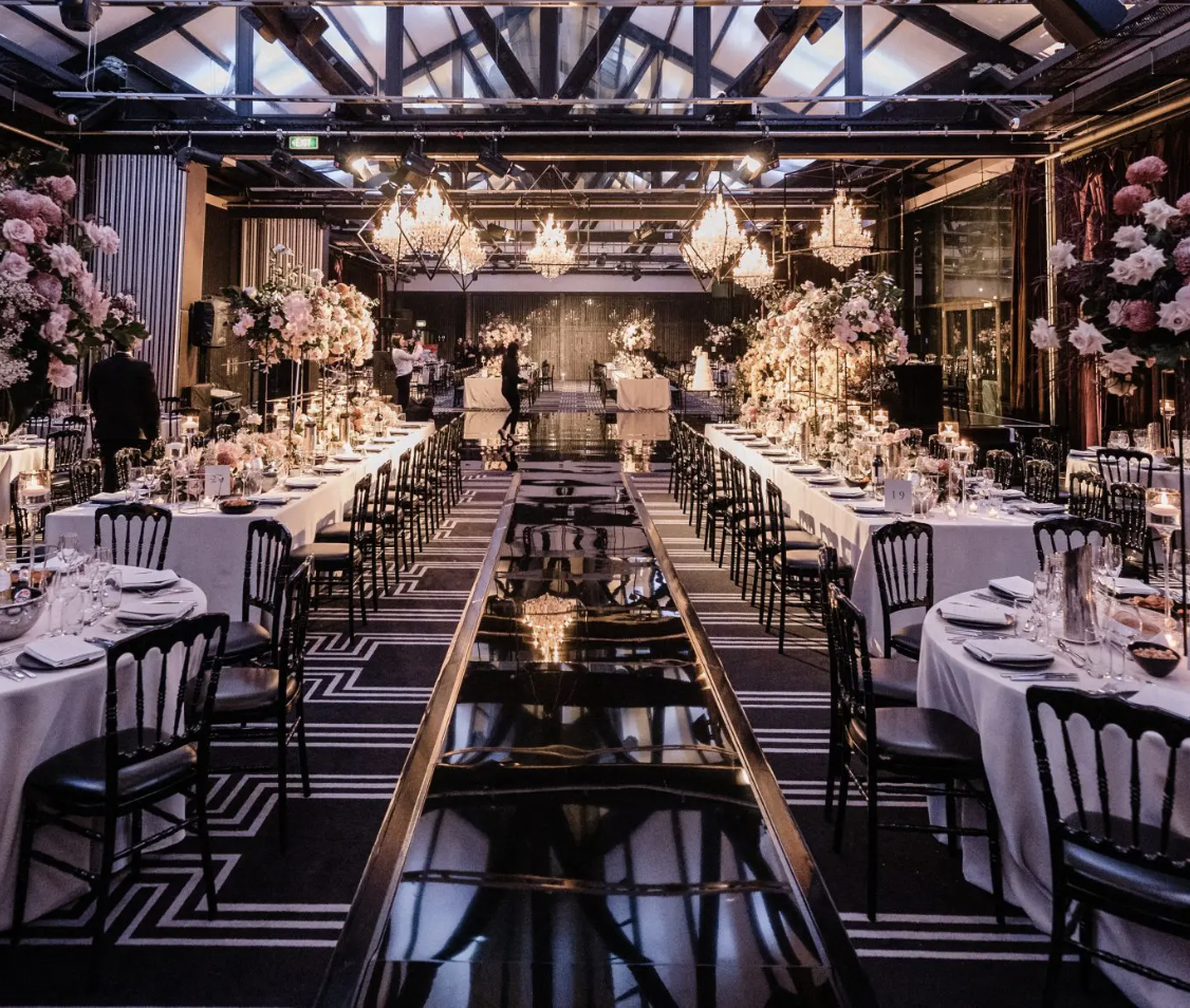 Elegant banquet hall decorated for a wedding reception with long tables covered in white tablecloths, tall flower arrangements with pink and white flowers, black chairs, and multiple chandeliers hanging from the ceiling.