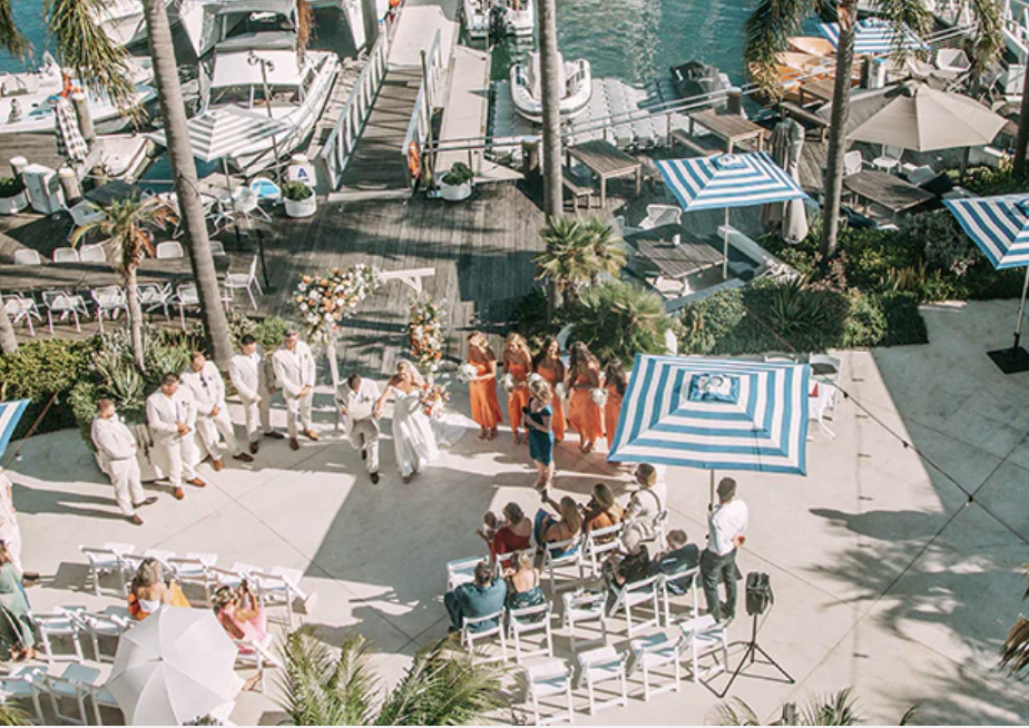 A wedding ceremony on a waterfront with guests seated, a floating dock with boats, and a couple walking down an outdoor aisle, surrounded by palm trees and umbrellas.