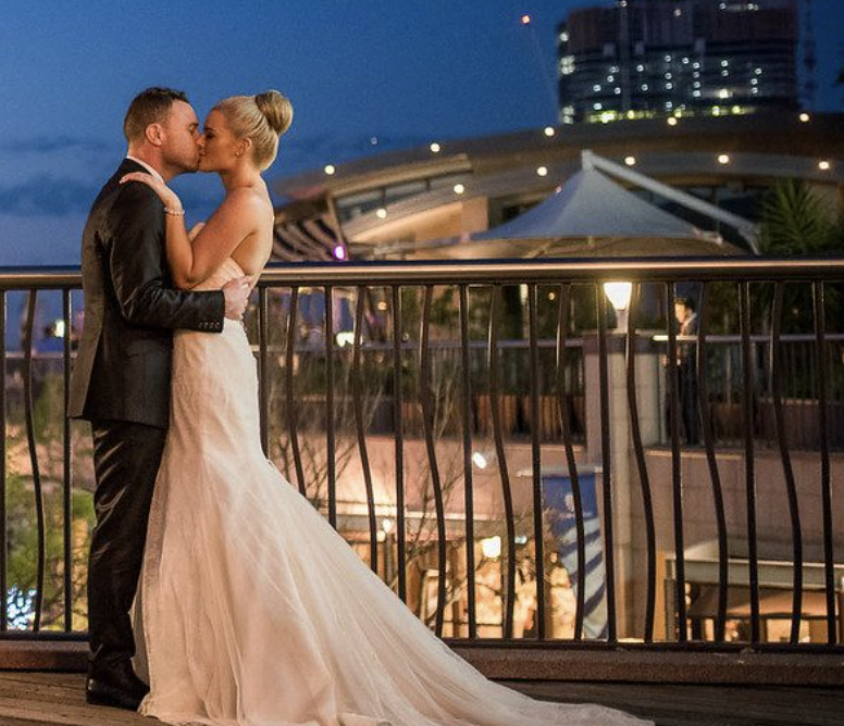 A bride and groom sharing a kiss on a balcony during sunset, with city buildings and a tent visible in the background.