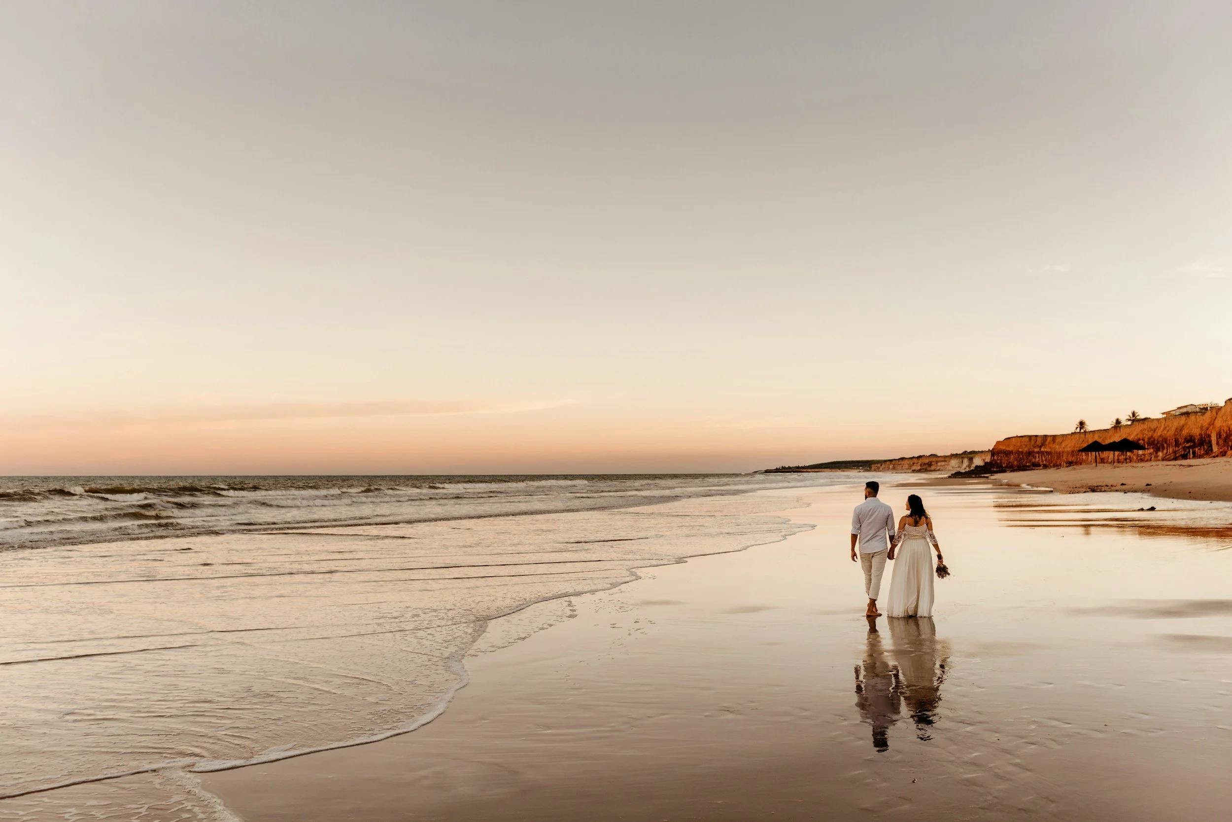 A couple walking along the beach at sunset, holding hands and carrying a bouquet, with cliffs and houses in the background.