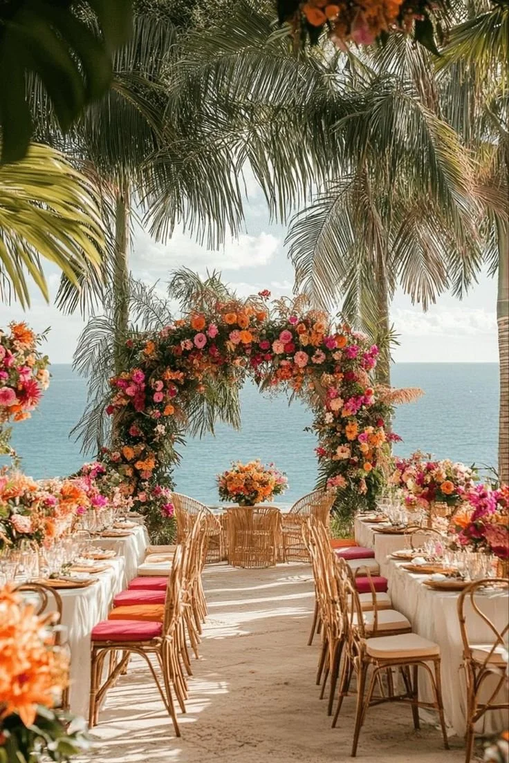 A tropical outdoor dining setup with long tables, surrounded by colorful flowers and palm trees, overlooking the ocean under a partly cloudy sky.