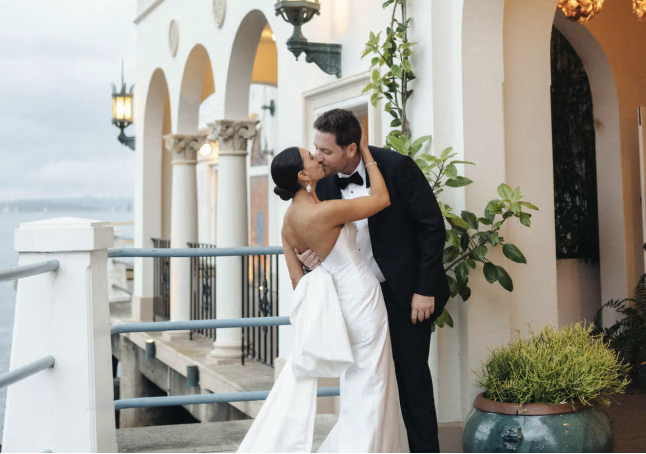 A bride and groom share a kiss on a balcony outside a building with arches, lanterns, and plants, during a wedding celebration.