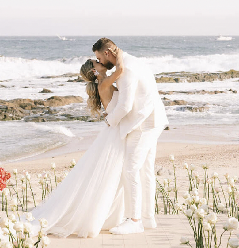 A couple in wedding attire sharing a kiss on a beach with flowers, rocks, and ocean waves in the background.