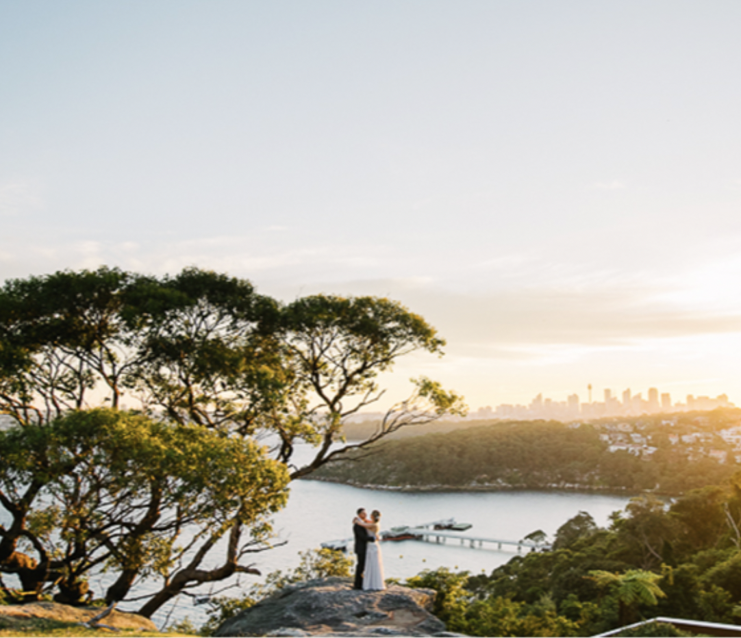 A couple standing on a rock overlooking a body of water with city skyline in the background during sunset.