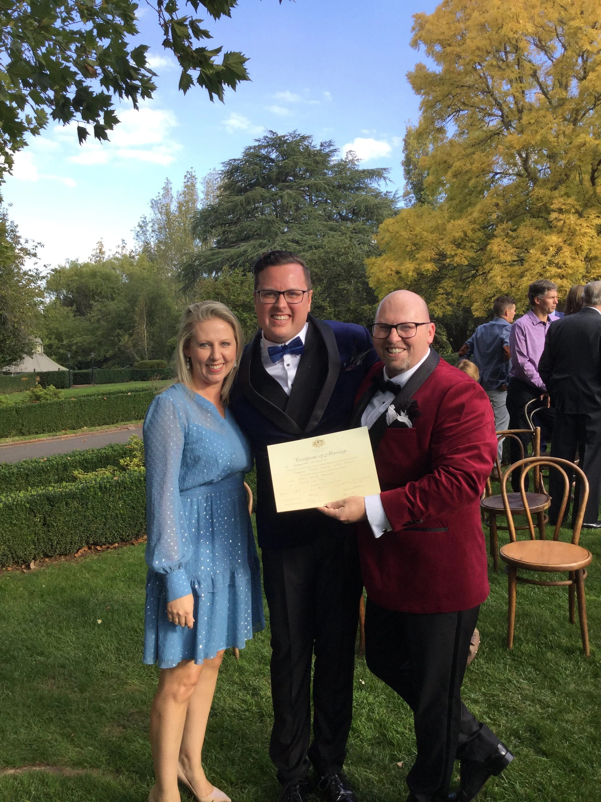 Three people celebrating at a wedding outdoors with trees and other guests in the background, holding a marriage certificate.