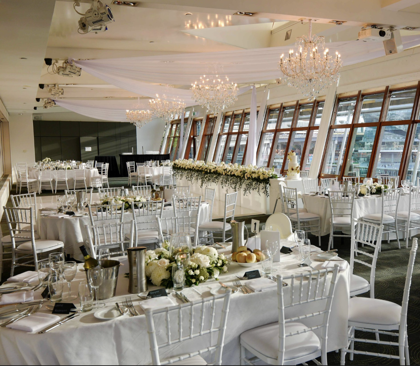 Elegant banquet hall decorated for a wedding reception with white tables and chairs, floral centerpieces, chandeliers, and large windows showing a harbor view.