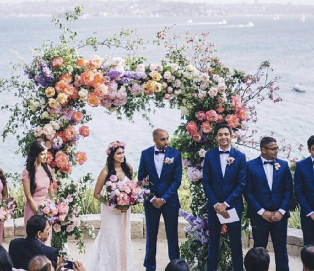A wedding ceremony with the bride and groom standing under a floral arch with pink, purple, and white flowers by the water. The bride holds a bouquet and wears a white dress; the groom and groomsmen wear blue tuxedos with black bow ties. Bridesmaids wear pink dresses. Guests are watching.