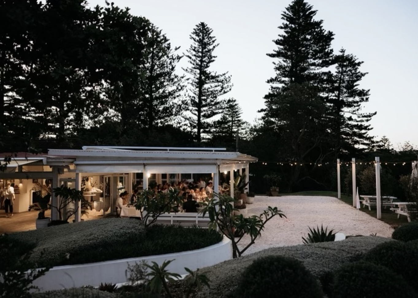 Dunes at Palm Beach's outdoor dining area at dusk with people sitting at tables under a covered patio, string lights overhead, and trees in the background.