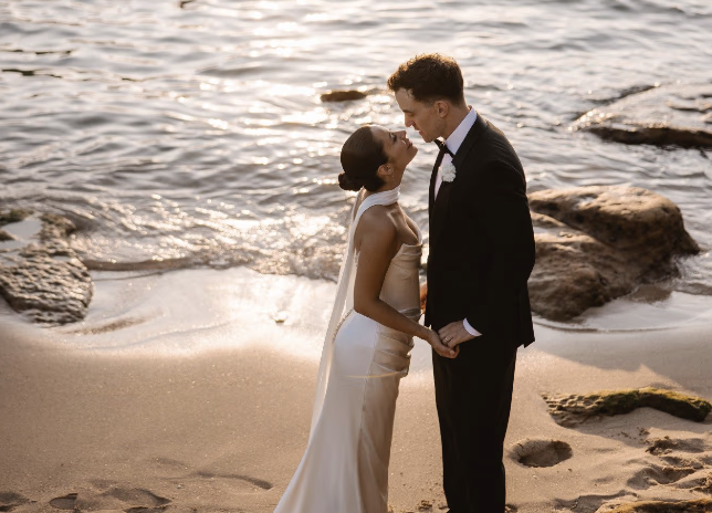 A bride and groom standing on a sandy beach, holding hands and gazing at each other at sunset with the ocean and rocks in the background.